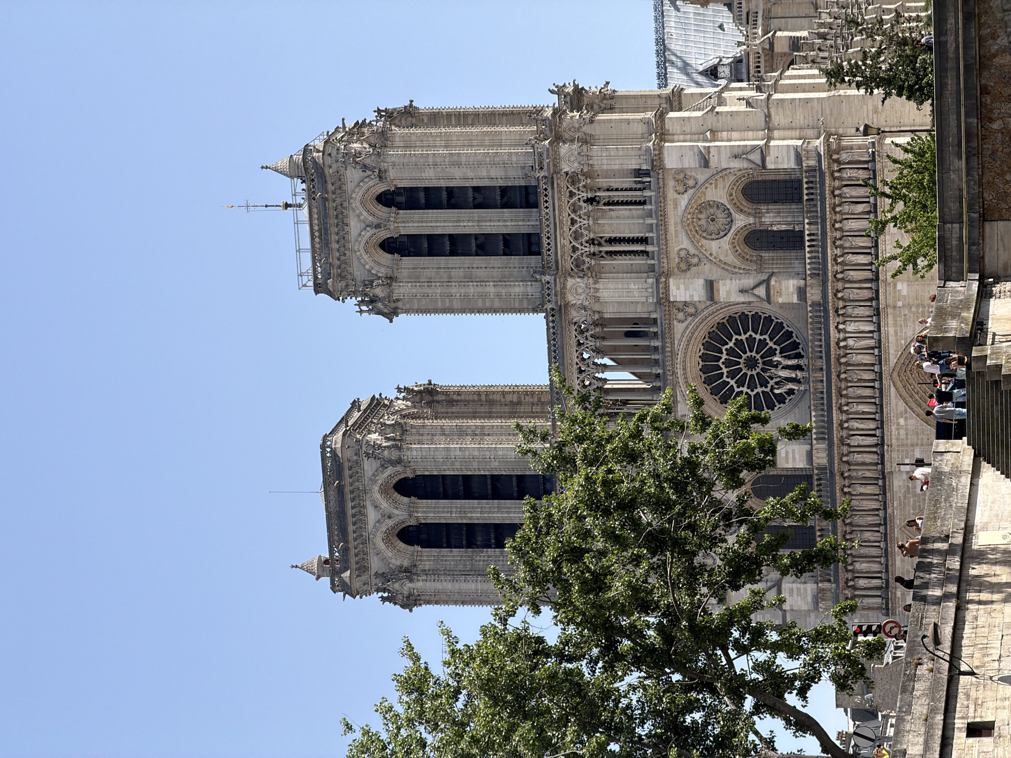 This image shows the Notre Dame Cathedral in Paris, France. The photo is taken from a low angle, emphasizing the towers and the western facade of the building. The architecture is in the Gothic style, featuring intricate stone carvings and a large rose window. Trees are visible in the foreground. People are standing along the exterior of the cathedral. The sky is light blue.