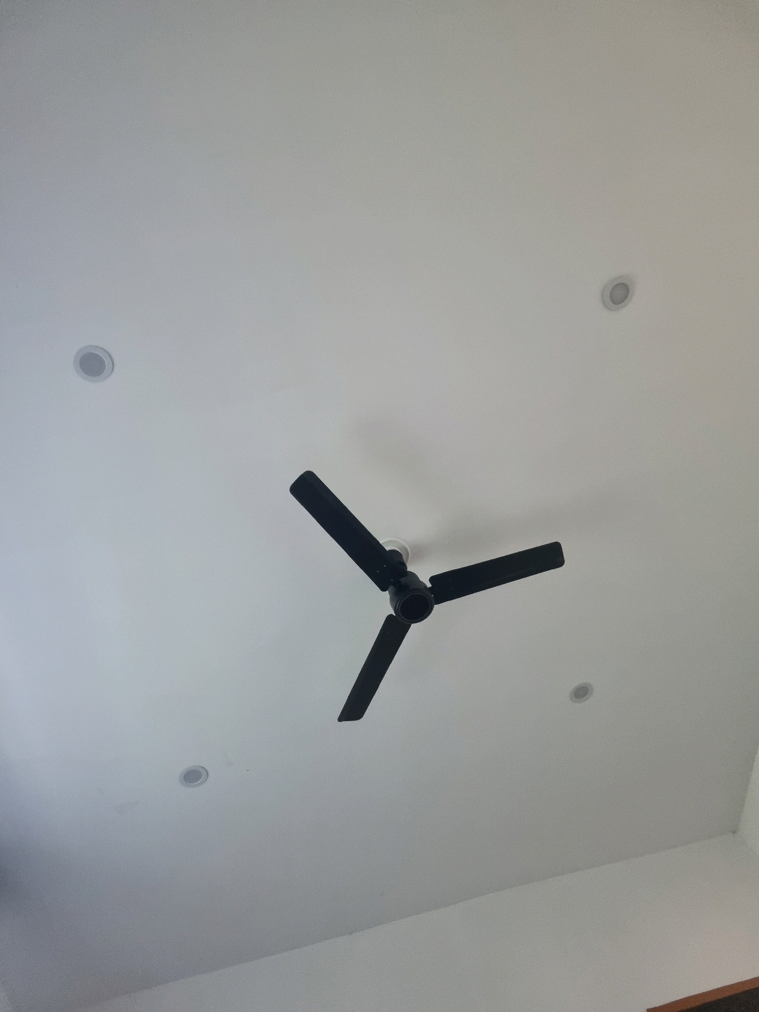 A black, three-bladed ceiling fan is suspended from a white ceiling, with its blades positioned at an angle. The ceiling also features four circular recessed lights, evenly spaced around the fan. The overall scene depicts an interior space, possibly a room in a residential or commercial building in Manigam, India. There are no people visible in the image.