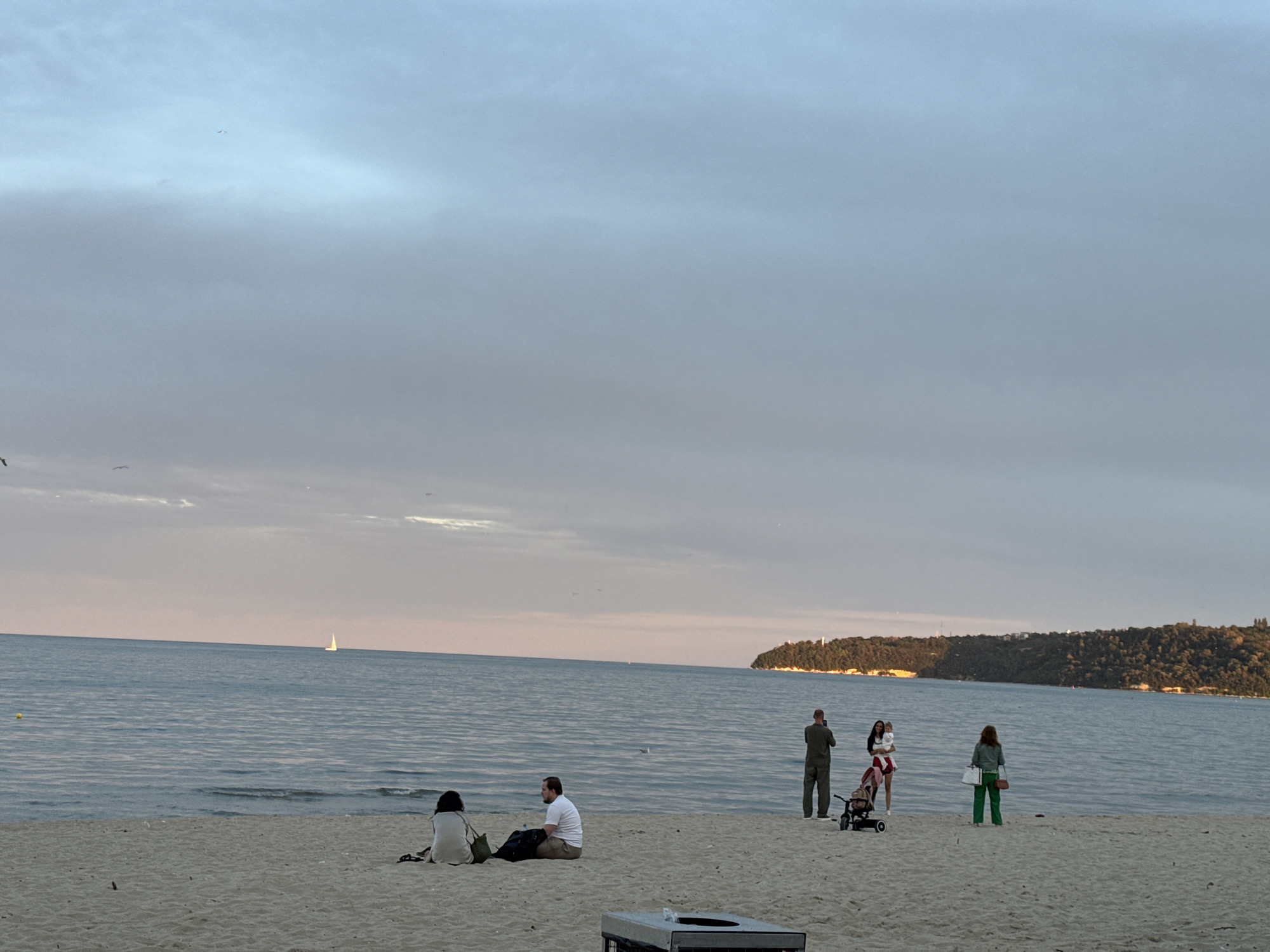 The image depicts a serene beach scene at sunset in Varna, Bulgaria. The sky is painted with soft pastel colors, with hues of pink and blue blending into the gentle clouds. The sea is calm, with small waves lapping at the sandy shore.
On the beach, several individuals are enjoying the evening. In the foreground, a couple sits together on the sand, facing the water. To their right, a group of three people stands near the water's edge. One person is holding a baby, while another person, possibly a parent, is standing nearby with a stroller. A fourth person, facing away from the camera, stands further to the right, holding a bag.
In the distance, a sailboat glides across the water, adding to the tranquil atmosphere. The coastline is lined with lush green trees, suggesting a natural landscape bordering the city. The overall impression is one of peaceful relaxation and natural beauty.