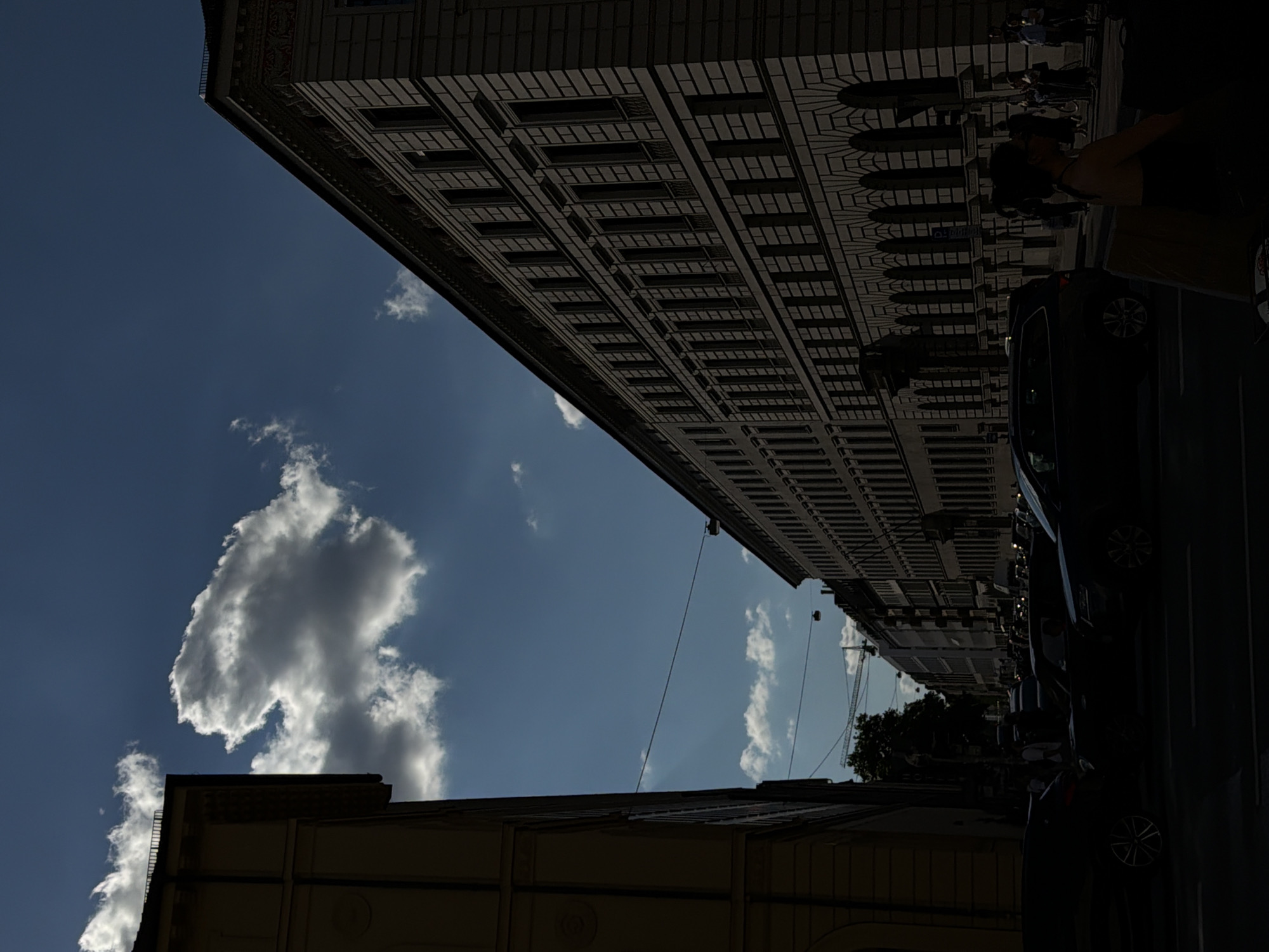 The image depicts a city street in Munich, Germany, characterized by architectural buildings and a street scene. On the left, a portion of a light-colored building with classical detailing is visible in shadow. Dominating the right side of the frame is a large, ornate building with multiple stories, visible in shadow. The sky above is a deep blue, with a prominent, bright white cloud and some smaller clouds scattered. Several cars are present on the street, rendered in deep shadow, indicating strong contrast between light and dark. In the foreground, a few figures are also in silhouette. A street light and overhead wires are visible, along with what appears to be a construction crane in the distance. The overall impression is a streetscape with a dramatic interplay of light and shadow, likely captured during a sunny day with overcast elements.