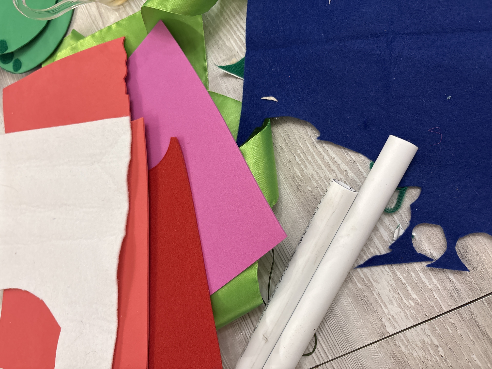 The image depicts a close-up, overhead view of various crafting materials spread across a light-colored, wooden surface. The materials include sheets of felt in red, pink, and blue, as well as green ribbon. There are also two white cylindrical objects, possibly PVC pipes, positioned diagonally across the lower left portion of the frame. In the upper right, partially visible, are some green circular shapes adorned with darker green dots, and a clear glass object with a handle, possibly a pitcher or vase. The wooden surface appears to be a tabletop or floor. The scene suggests a crafting or DIY project in progress. There is no discernible information within the image that would allow for the determination of its location in the city of Izhevsk, Russia.