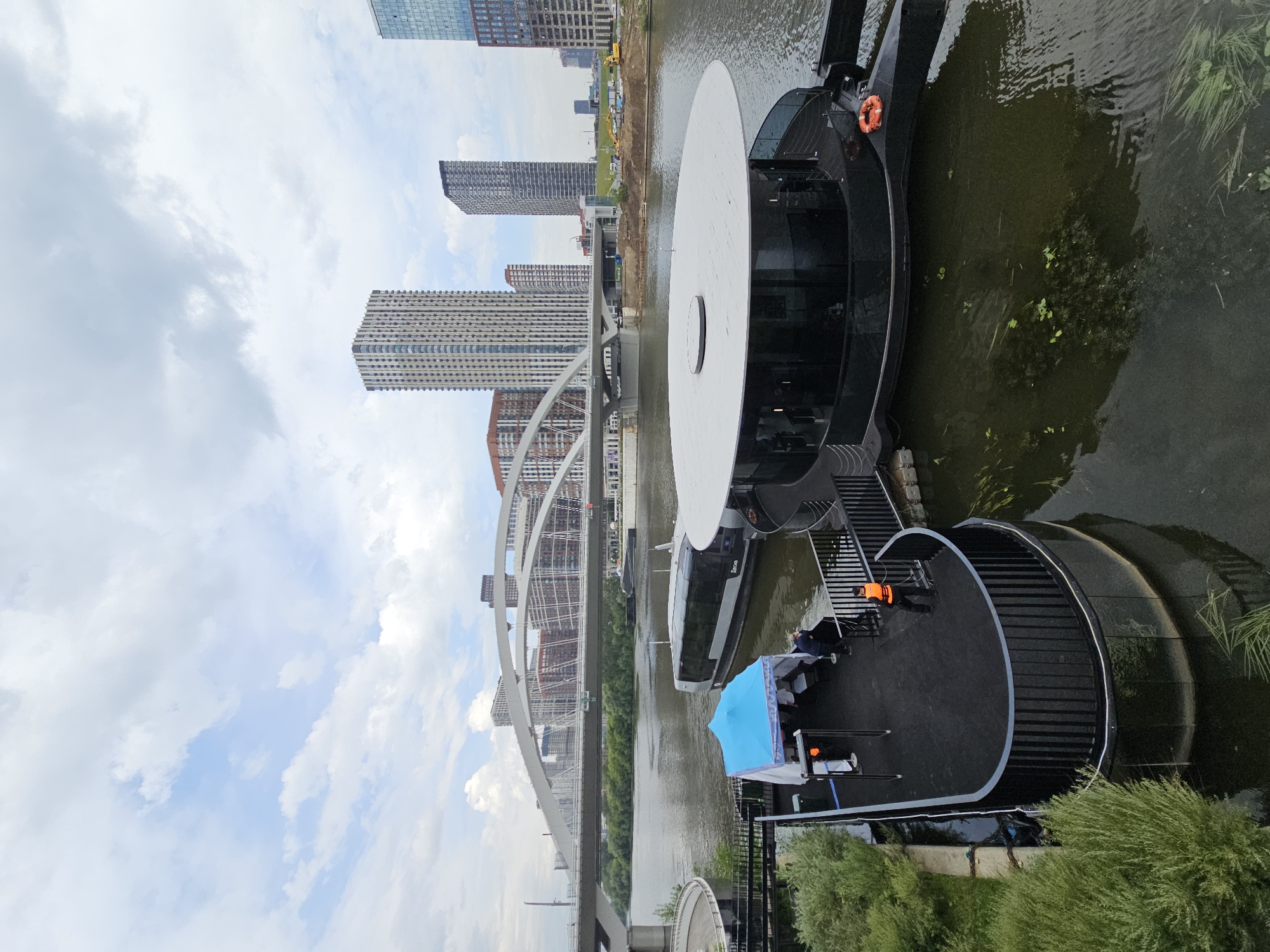 A wide-angle, eye-level shot captures a modern, circular, black-hulled boat docked on a calm river in Moscow, Russia, under a partly cloudy sky. The boat features a large, white, flat circular roof and large glass windows. Adjacent to the boat, a smaller, curved platform is also docked, covered by a blue and white tent, under which several people are standing. A person in an orange life vest stands on the edge of the platform, facing away from the camera. In the background, a large, white arched bridge spans the river, with numerous tall, modern apartment buildings and skyscrapers visible beyond it. The riverbanks are lined with green trees and shrubs. Construction equipment, including excavators, is visible on the far riverbank. The water is murky and shows some submerged vegetation. The overall scene suggests a contemporary urban waterfront area.