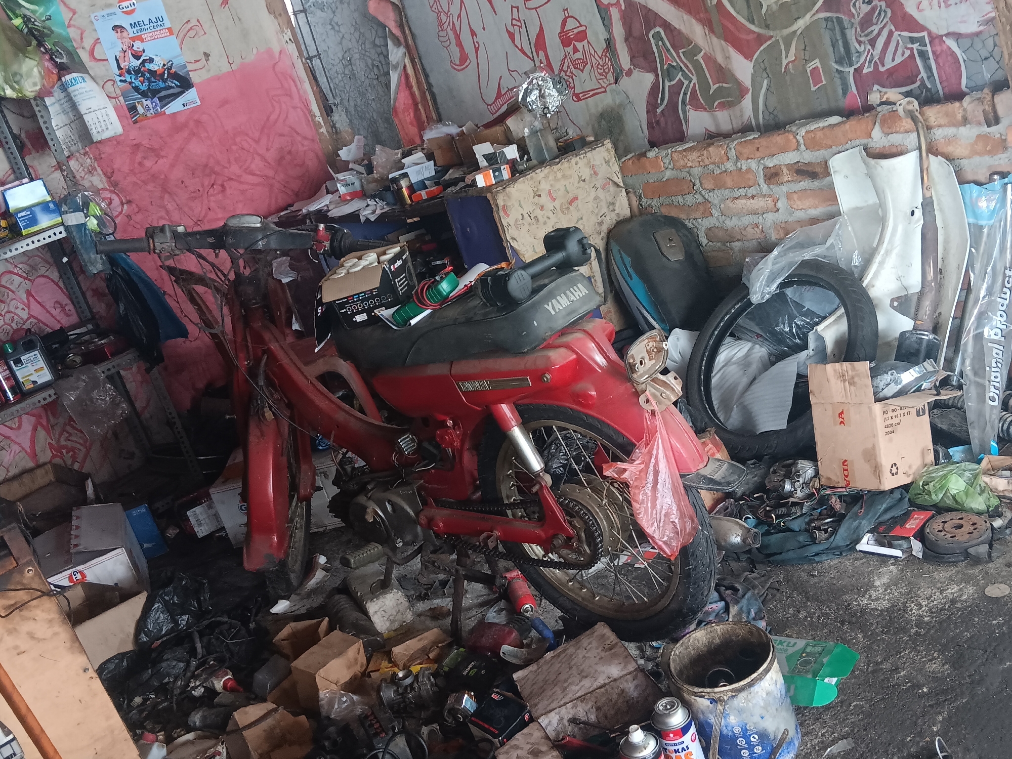 The image depicts a cluttered motorcycle repair shop, likely in Tlogo, Indonesia. The scene is filled with various motorcycle parts, tools, and debris. A red motorcycle is partially visible in the foreground, with its engine and wheels exposed, suggesting it is undergoing repair. Various boxes, plastic bags, and cans are scattered around, along with a toolbox and other mechanical components. The walls are adorned with posters and graffiti, adding to the workshop's chaotic yet functional atmosphere. The overall impression is one of a busy and active repair environment.