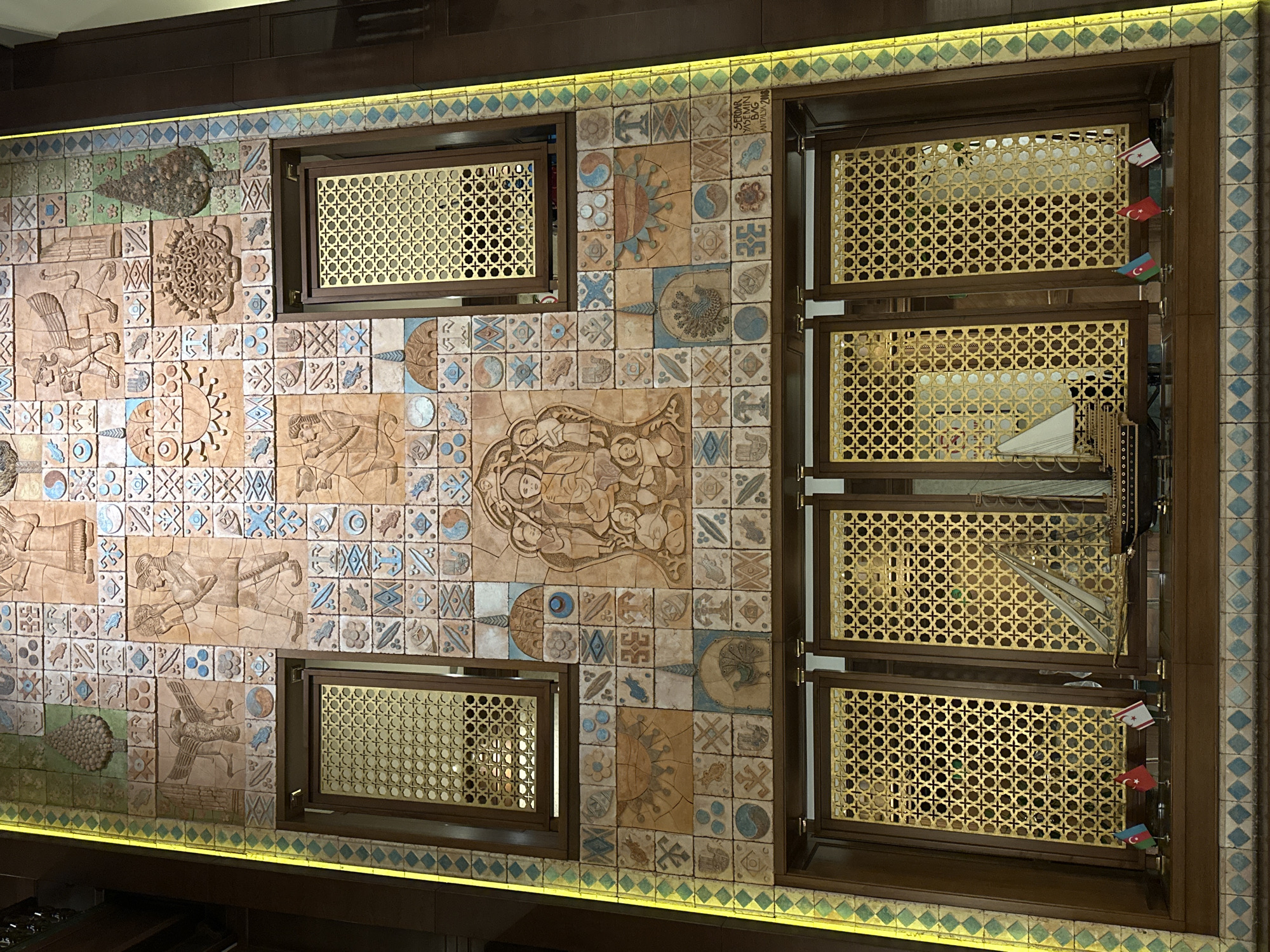 The image shows a large, ornate tile mosaic wall in Ankara, Turkey, likely a decorative element in a public building or establishment. The mosaic is a complex tapestry of small, square tiles featuring various embossed designs and patterns. Central to the mosaic is a depiction of a seated figure with two smaller figures, possibly a religious or historical scene. Surrounding this central motif are numerous smaller tiles, many with geometric patterns, stylized animals (such as birds and mythical creatures), celestial bodies (sun-like motifs), and symbolic shapes.

The mosaic is framed by wooden elements and incorporates two rectangular windows with decorative gold latticework. Below these windows, a lower section also features gold latticework behind which miniature ship models are displayed. Three small flags stand on a shelf in front of these models: the flag of Azerbaijan, the flag of Turkey, and the flag of the Turkish Republic of Northern Cyprus.

The overall impression is one of rich cultural and artistic expression, likely intended to convey historical or symbolic meaning. The presence of the flags suggests a connection to Azerbaijani and Turkish heritage, possibly indicating the location of an embassy, cultural center, or a business with strong ties to these regions. The mosaic's intricate detail and prominent display suggest it is a significant artistic feature of the space.