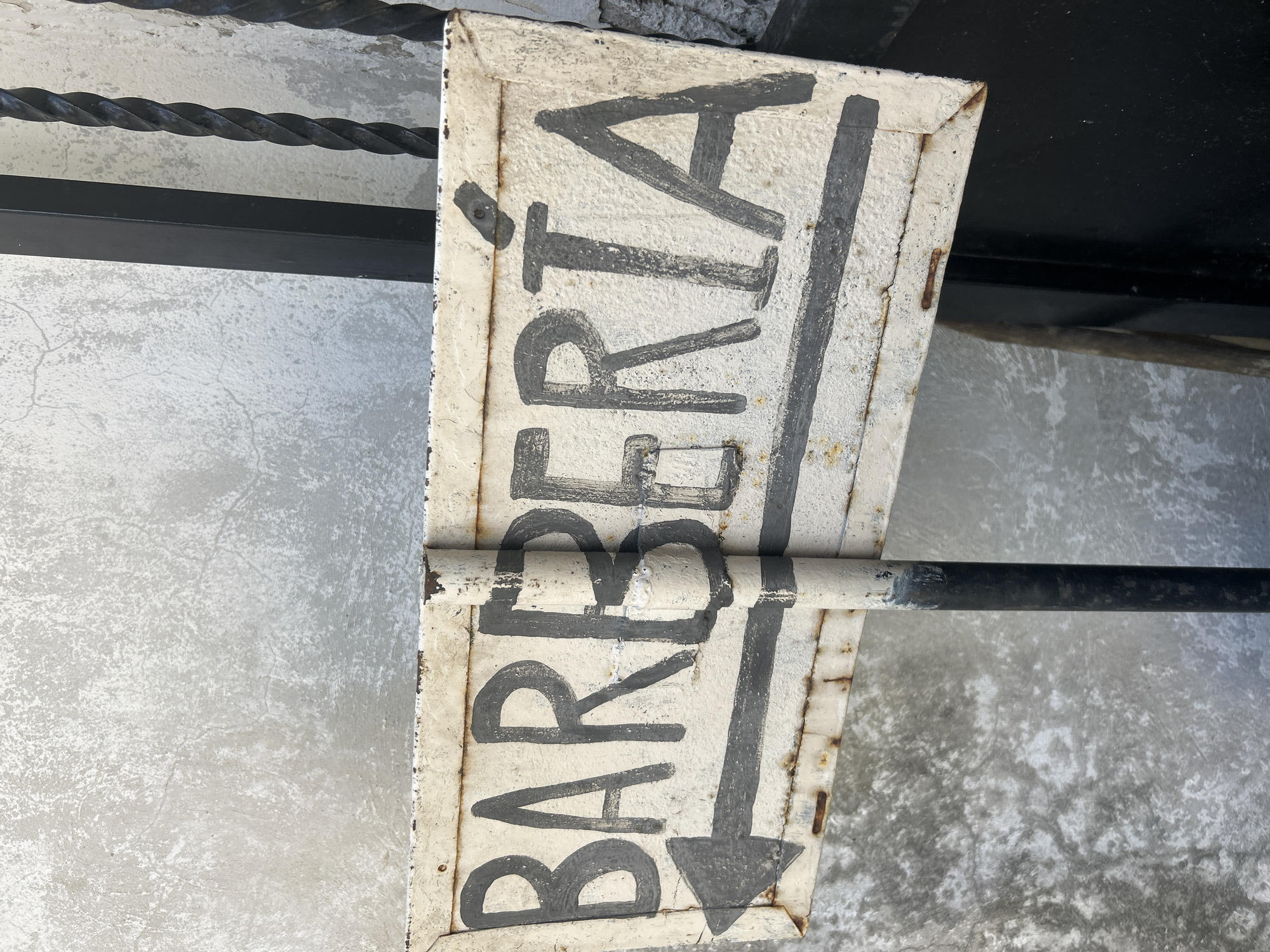 A weathered, off-white metal sign is mounted on a metal pole. The sign has a rectangular shape and is oriented horizontally. Bold, dark gray lettering spells out "BARBERÍA" across the sign, with an arrow pointing to the left above a horizontal line also in dark gray. The sign shows signs of rust and wear, particularly along the edges and in the paint. The background consists of a textured, light gray concrete wall on the left and black metal bars and a black surface on the right. The sign appears to be an outdoor advertisement or directional marker. The scene is consistent with a street-level location in the city of Santa Ana, Venezuela, likely indicating the presence of a barbershop in the direction of the arrow.
