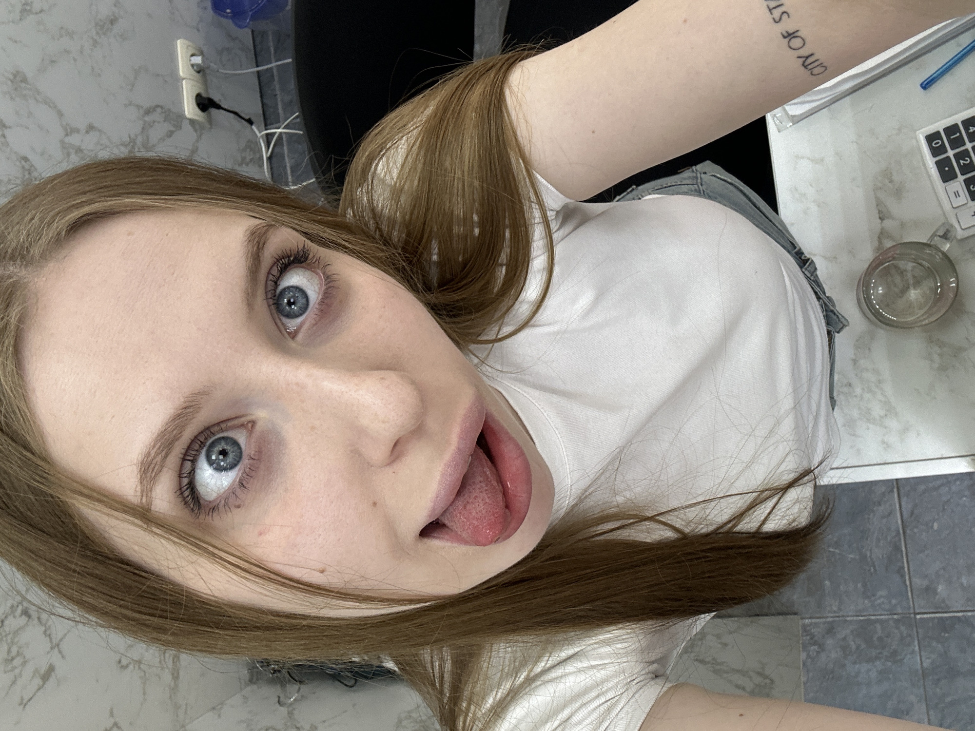 A young woman with light brown hair and blue eyes is taking a selfie. She is wearing a white t-shirt and what appears to be denim shorts. Her tongue is sticking out slightly. To the right of her, there is a desk or countertop with a calculator, a pen, and a glass mug. The background includes a marble-patterned surface and an electrical outlet. There is no information to determine that the location is in the city of Chaikovsky, Russia.