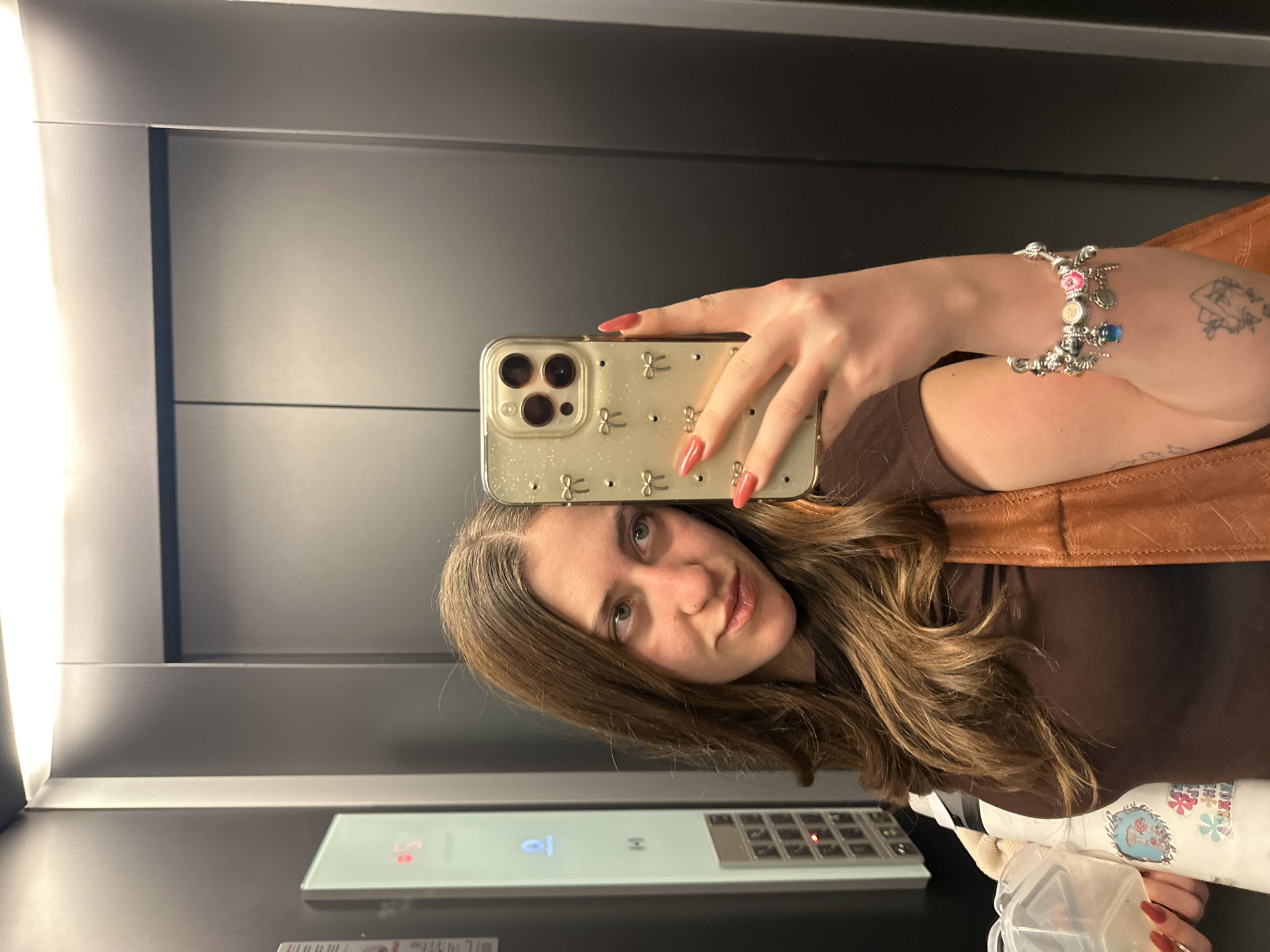 A person is taking a selfie in an elevator. The person has brown hair, fair skin, and is wearing a brown shirt and a brown leather vest. They are holding a gold iPhone with a decorative case. The person is wearing a charm bracelet on their left wrist. The elevator has gray walls and a panel with buttons. A water bottle with stickers is also visible. The location is not discernible from the image alone and therefore cannot be confirmed as Huixquilucan de Degollado, Mexico.