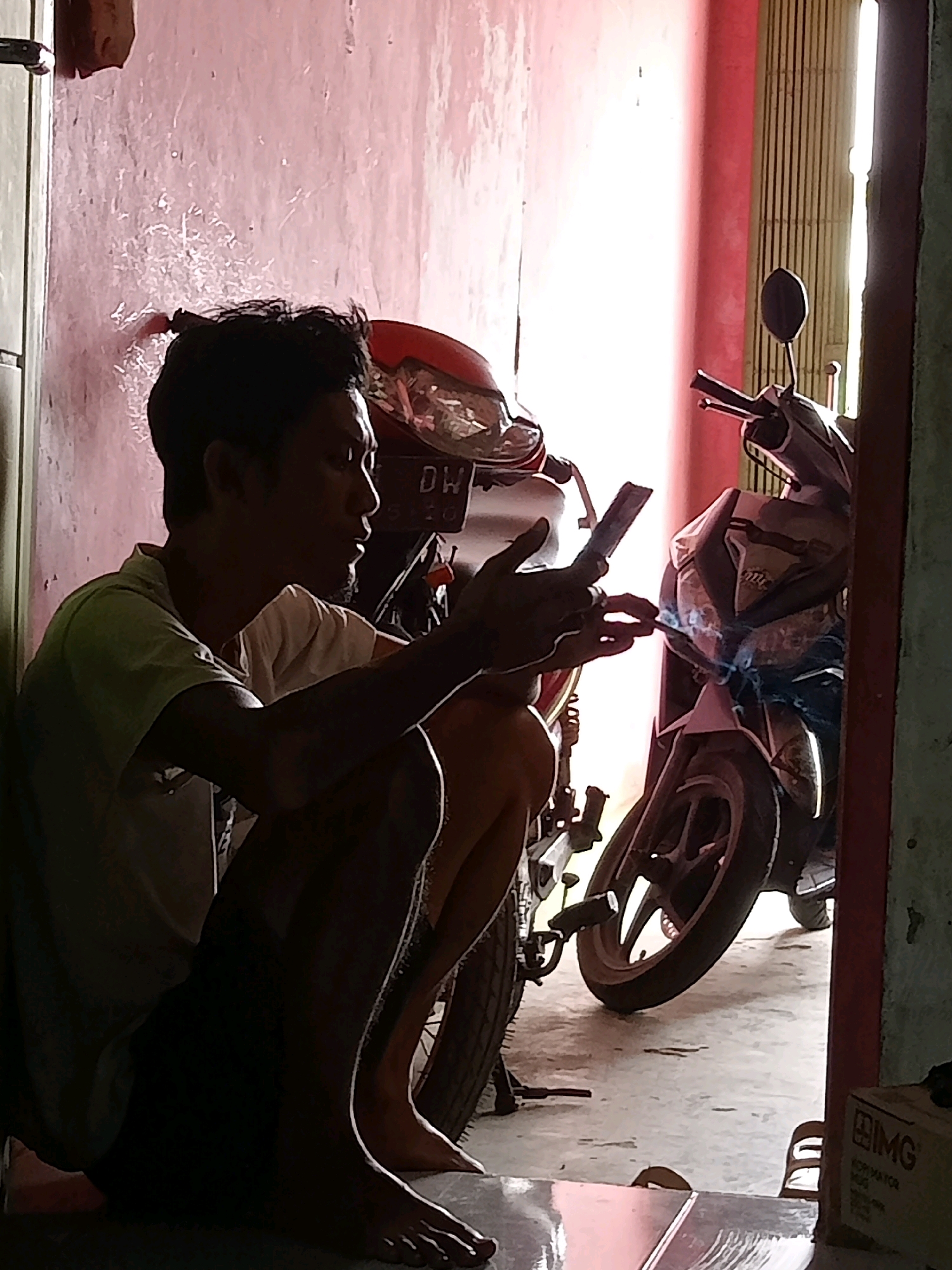 A man is sitting on the ground, silhouetted against a bright pink wall in what appears to be an open doorway or alleyway. He is wearing a light-colored t-shirt and dark shorts, with his legs crossed. He is holding a smartphone in his right hand and appears to be looking at it. In front of him, to his right, is a motorcycle with a silver and black exterior. Sparks are visible between the man's left hand and the motorcycle, suggesting he is welding or performing some sort of repair. The floor is concrete, and the background shows a closed metal gate or shutter. The scene is dimly lit, with a strong light source coming from the right side of the image, creating harsh shadows. The overall impression is one of a skilled worker engaged in a task.