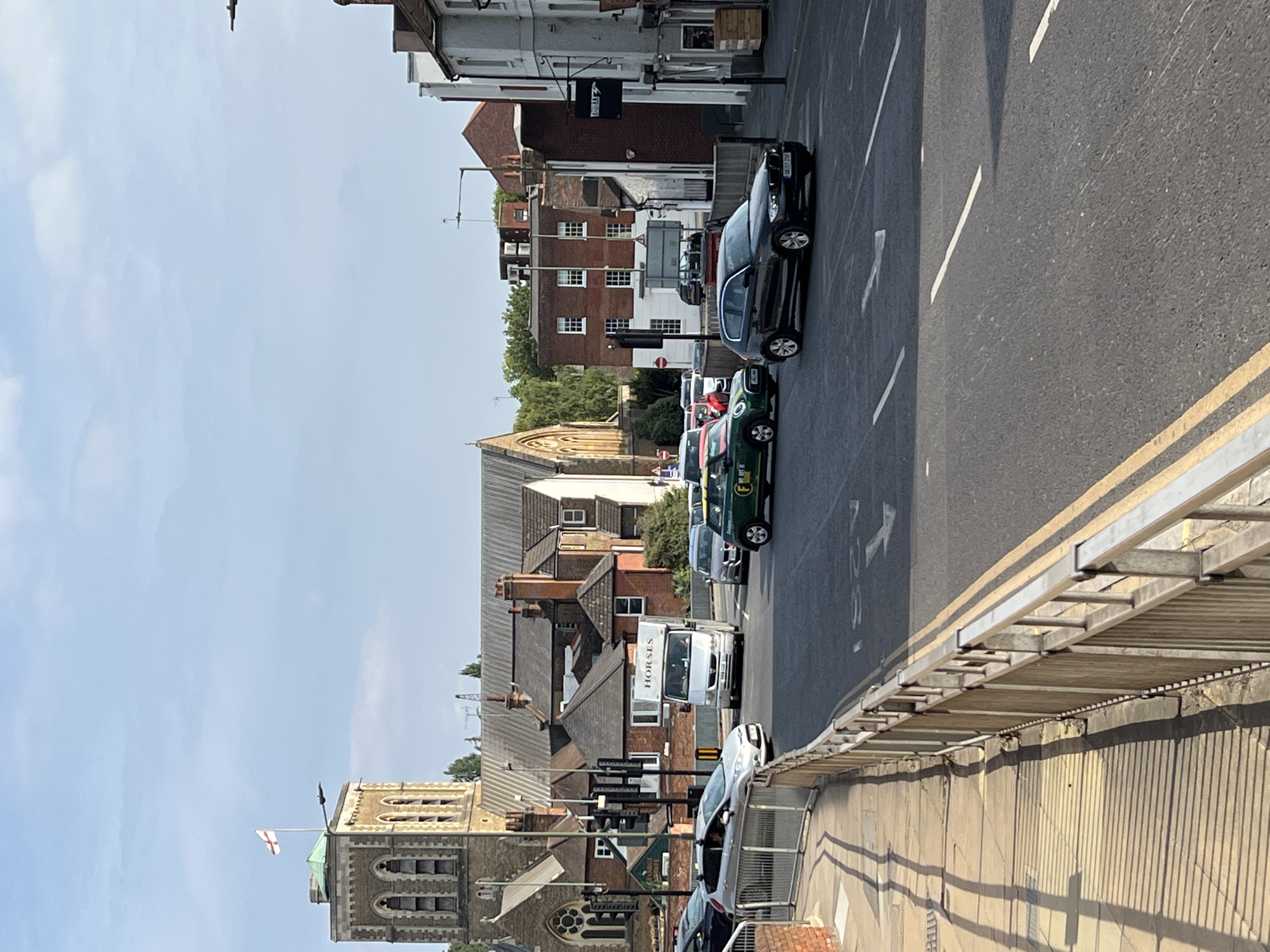 This image captures a street scene in Guildford, United Kingdom, on a bright, sunny day. The perspective is from an elevated viewpoint, looking down a street lined with buildings and a road.
The foreground features a metal railing and a concrete structure, casting long shadows. Beyond this, a road is visible with yellow and white lane markings, indicating a street in use. Several cars are parked and in motion on the road, including a dark-colored sedan, a green Mini Cooper, and a white van with "HORSES" written on its side. A silver car is also seen on the right side of the road.
The buildings along the street vary in architecture. Some appear to be older brick structures with multiple windows, while others are more modern in design. A prominent building with a bell tower, likely a church, is visible on the left side of the image, flying an English flag. Another building with a distinctive arched entrance is also present.
The sky is clear and blue, suggesting a pleasant time of day, likely midday or early afternoon. The overall atmosphere appears to be calm and ordinary, depicting a typical day in a town. There are no specific events or activities clearly discernible, other than the movement of traffic and pedestrians (implied by the parked cars and buildings).
Visible text includes the word "HORSES" on the white van and the license plate of the dark sedan, which appears to read "BD09 JRX". Additionally, a sign on the road has an arrow and the number "28". A black sign with white lettering hangs from a pole in front of one of the buildings, but the text is too small to read.