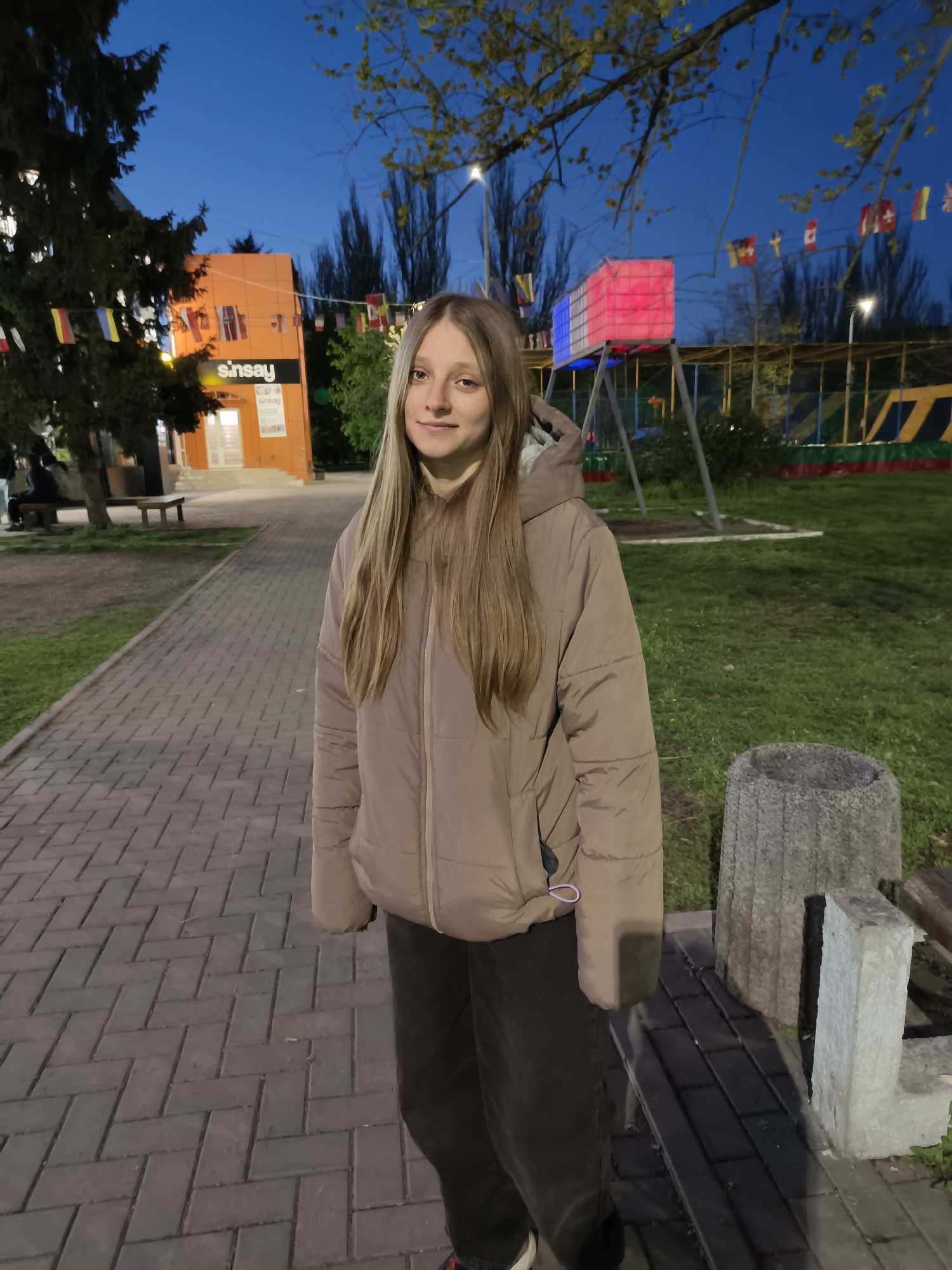 The image depicts a young woman standing on a paved walkway in an outdoor public space during the early evening in Kryvyi Rih, Ukraine. The sky is a deep twilight blue, and artificial lights illuminate the scene.
The main subject is a young woman, likely a teenager or young adult, with long, straight, light brown hair. She is wearing a beige or light brown puffer jacket and dark pants, and she smiles faintly while looking directly at the camera.
In the background to her left, an orange building features two white signs displaying the text "sinsay." A white door is visible beneath the signs. A large, dark green tree partially obscures this building. To the right and further back, a recreational area includes a prominent rectangular light structure illuminated in red, white, and blue sections. Below it, parts of a colorful playground or trampoline area with green and red details are visible. A string of various national flags, including those of Canada, Switzerland, and the United Kingdom, is strung across the background. Streetlights are also visible throughout the scene.
The foreground features a textured brick paved path where the woman stands. To the right of the path, a gray stone trash can and another stone block are present. The overall setting appears to be a park or town square, bustling with decorative elements suggesting a lively, community-oriented atmosphere.