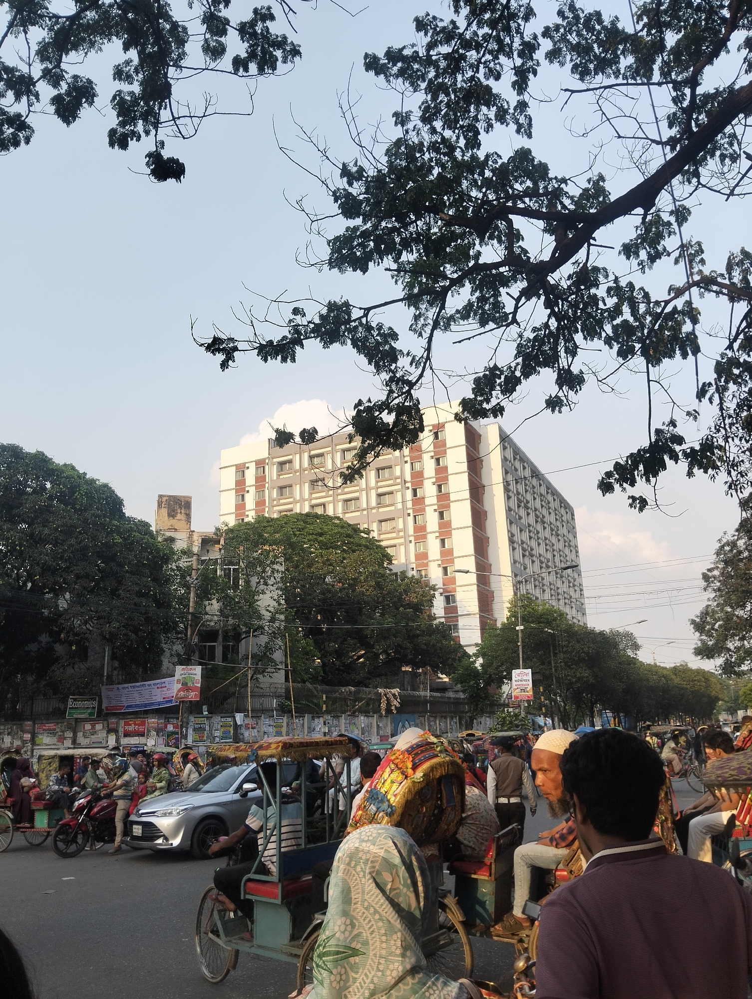 The image depicts a busy street scene in Dhaka, Bangladesh. Numerous rickshaws, motorcycles, and a silver sedan are visible amid pedestrians. A tall, rectangular building with a white and grey facade, accented with vertical red stripes, stands in the background. Trees partially obscure the building, with tree branches also present in the upper portion of the frame. The sky is a light blue, almost white. People are densely packed near the intersection, creating a bustling atmosphere. Posters and signs are affixed to buildings along the roadside.