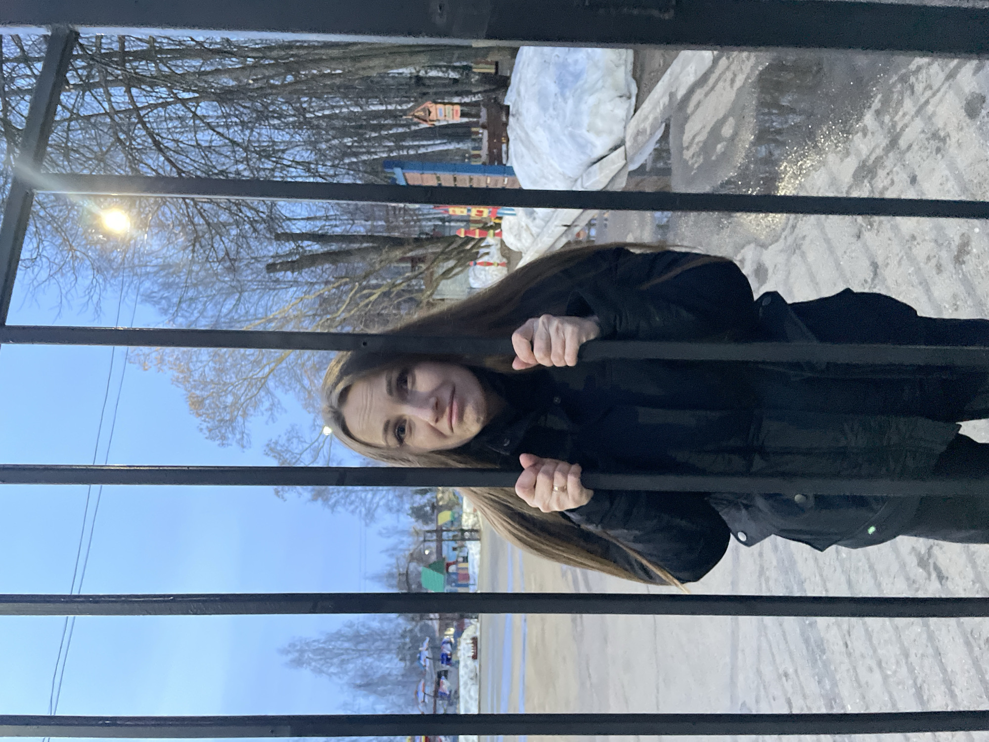 A medium shot shows a Caucasian woman standing behind a dark metal fence, looking toward the viewer. She is wearing a dark jacket and has long, light-brown hair. Her hands grip the fence. Through the fence, one can see a winter scene with bare trees, snow-covered ground, and playground equipment. This photograph was taken in the city of Chaikovsky, Russia.