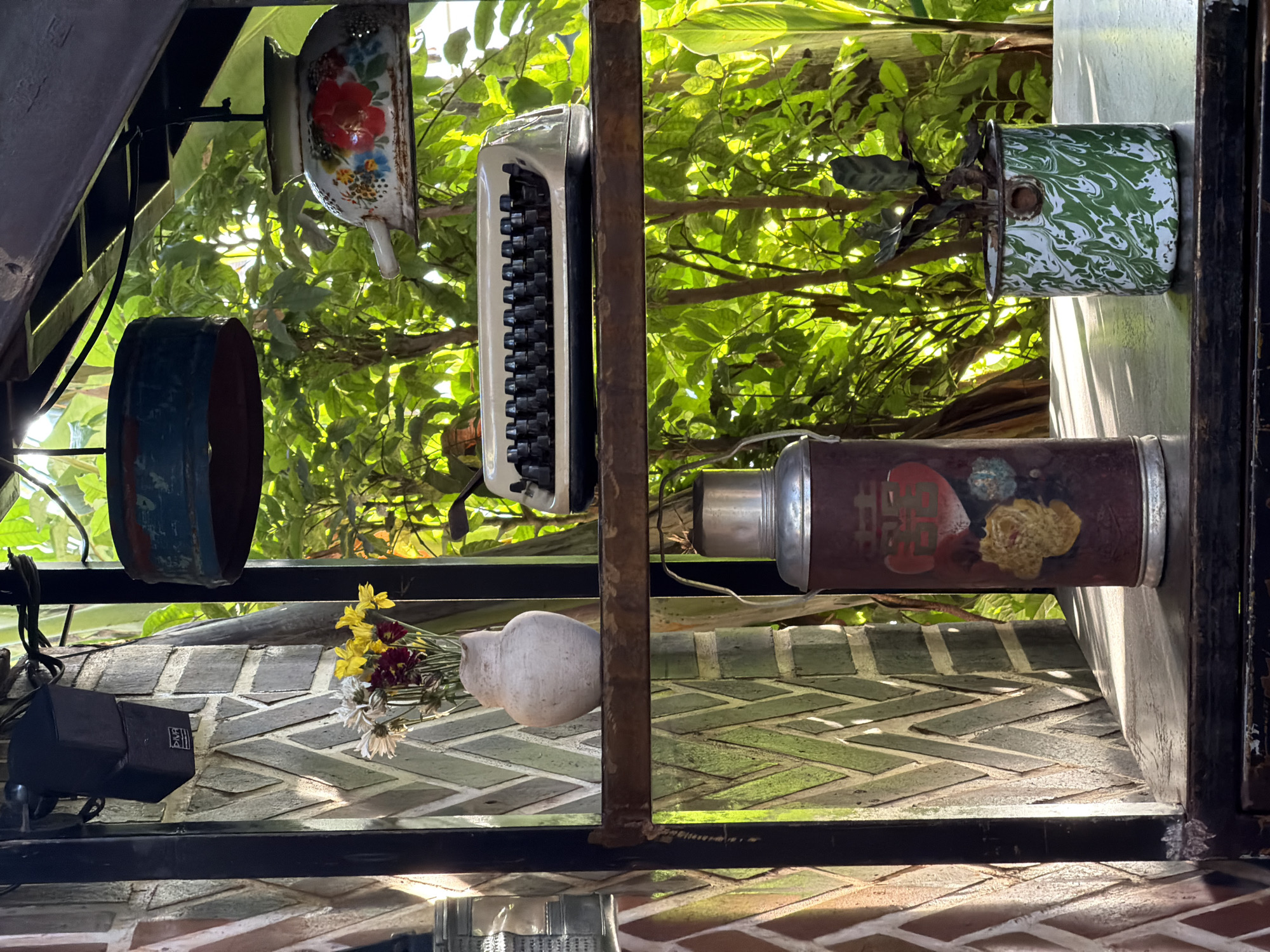 This image captures a shelf unit in what appears to be an outdoor or semi-outdoor setting, possibly a cafe or restaurant in Kuta Selatan, Indonesia, given the vibrant greenery and natural light. On the top shelf, a vintage white typewriter sits centrally, framed by lush green foliage. Above this shelf, two decorative hanging lamps are visible, one with a blue and red rim and the other a white enameled pot adorned with floral motifs. On the lower shelf, a large, cylindrical thermos with a decorative design is positioned to the left. To its right, a small potted plant in a green and white marbled enamel container adds a touch of life. A brick wall with a distinctive chevron pattern forms the backdrop on the left side of the image, suggesting an architectural element. The overall scene conveys a sense of rustic charm and a collection of vintage items displayed against a natural, sun-dappled environment.