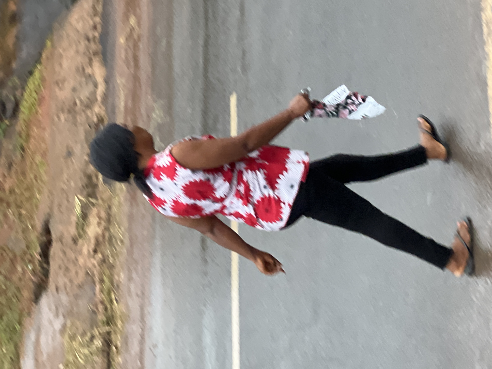 A woman, viewed from the side, walks on a wet asphalt road. She has dark hair pulled back and wears a sleeveless red and white floral print top and black pants. In her right hand, she carries a folded white and red patterned cloth. She is wearing black flip-flops. The road appears wet, possibly from rain, with a faint yellow line visible across it. The background is blurred, showing patches of grass and soil. The scene is set in Nkpor, Nigeria, on what appears to be a drizzly day.