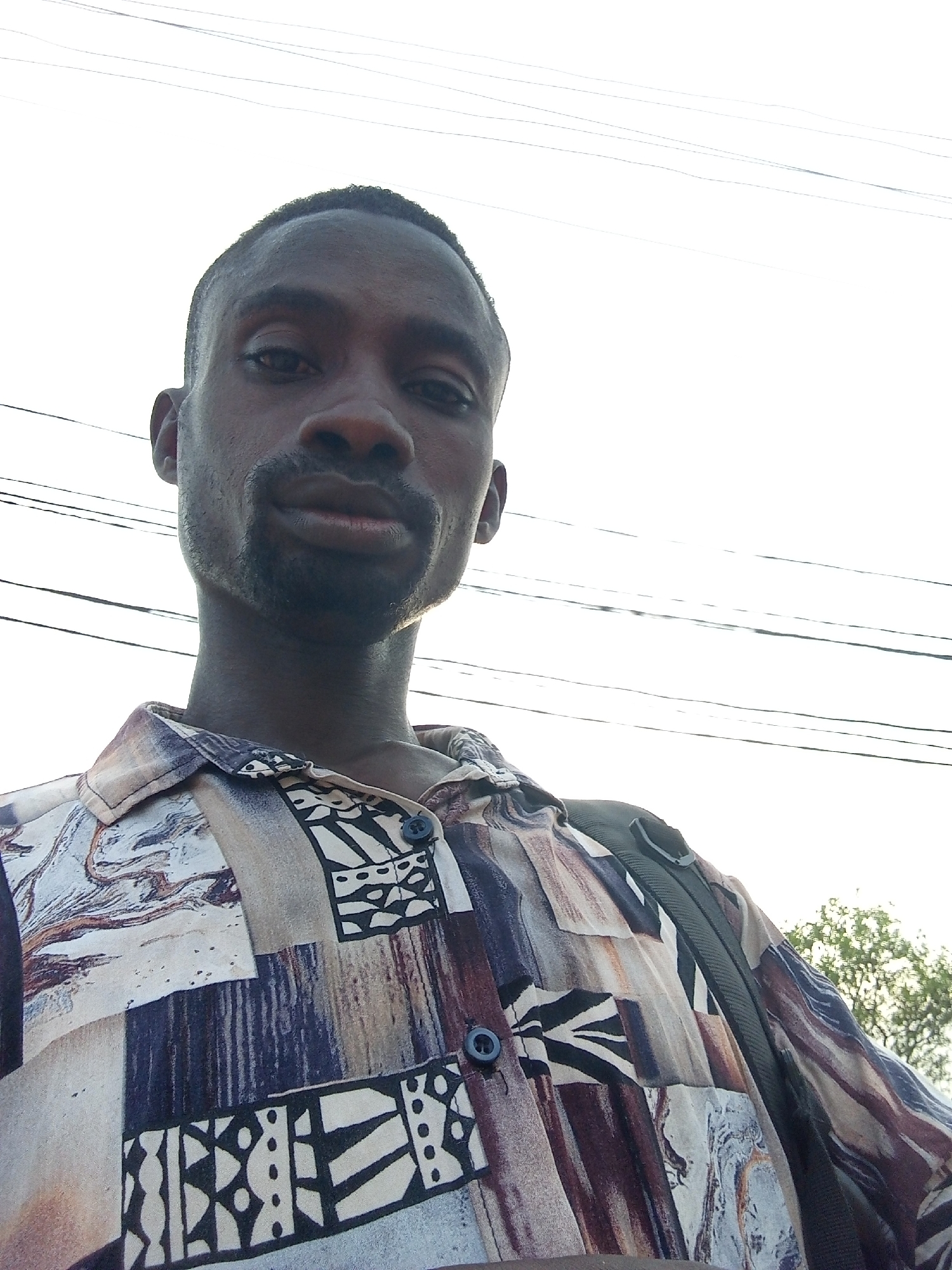 A man is standing outdoors, looking directly at the camera. He has dark skin, a close-cropped haircut, and a short beard. He is wearing a patterned button-down shirt. A black strap, possibly from a bag, is visible over his shoulder. Overhead, there are electrical wires against a bright sky. A tree is visible in the lower right background. The image was most likely taken during the day in Lokoja, Nigeria.