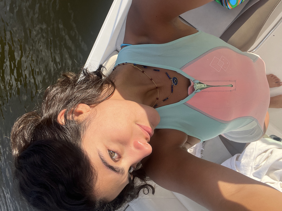 A young adult female is depicted in a close-up, high-angle shot aboard a white boat. She has dark, wavy hair and light-colored eyes. Her complexion appears tanned, with direct sunlight illuminating her face and casting shadows. She wears a sleeveless, two-tone garment, likely a wetsuit or rashguard, featuring a light blue upper section and a light pink main body with a central zipper and a subtle geometric logo. Two layered necklaces are visible: a gold chain with small red beads and a longer necklace with blue beads and an oval blue pendant. The subject is looking towards the camera. Behind her, calm, dark water is visible, indicating the boat is on a river or lake. This scene occurs on the Minho River, adjacent to the city of Vila Nova de Cerveira, Portugal.