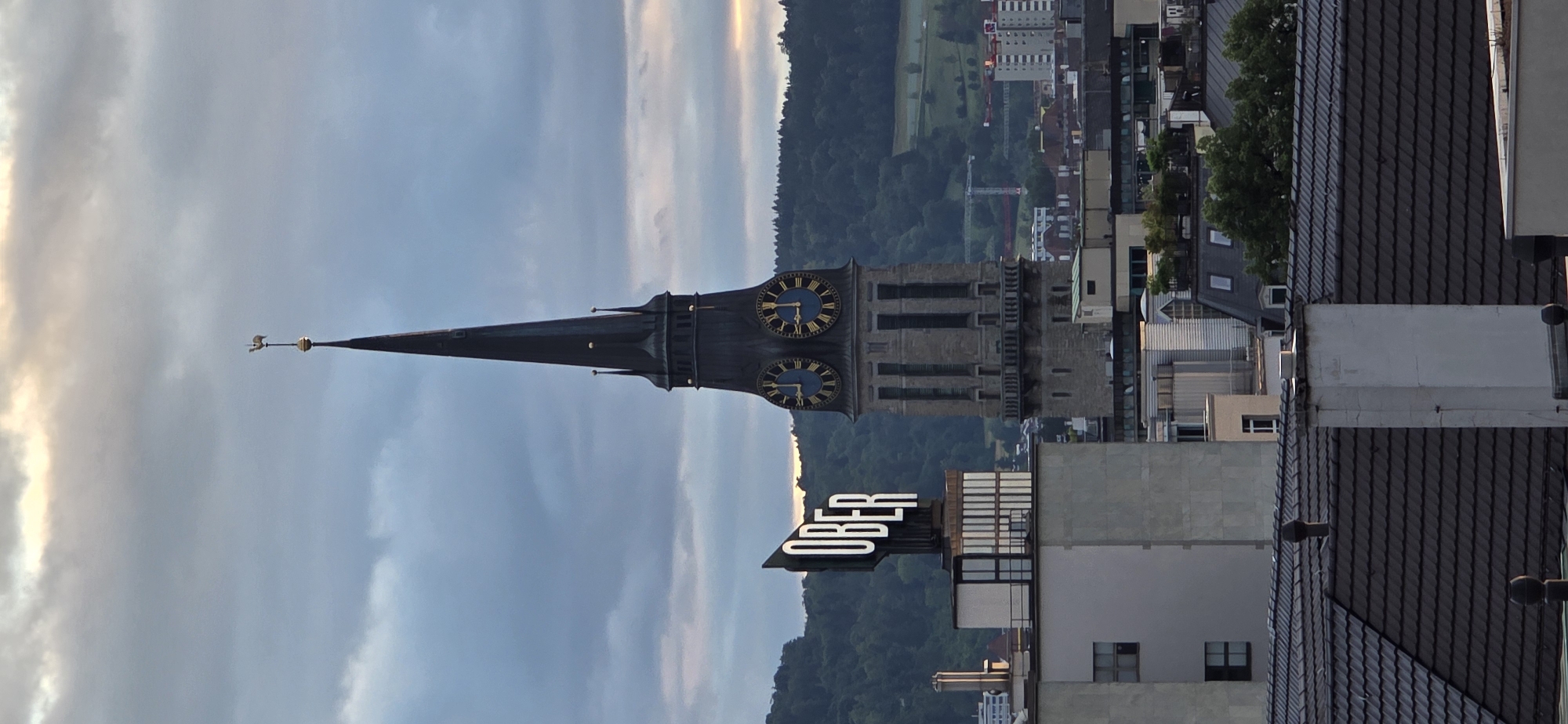 A twilight cityscape features a tall, dark, Gothic-style church spire with a weather vane at its apex. The spire has two prominent clock faces with Roman numerals and blue backgrounds. To the left of the spire, a rectangular building with the white illuminated word "OBER" on its roof is visible. The cityscape is comprised of various buildings of different heights and architectural styles, with a background of lush green hills. The sky is overcast with dramatic clouds, hinting at the end of the day. The foreground shows the dark, tiled roofs of residential buildings. The overall impression is one of a European city at dusk.