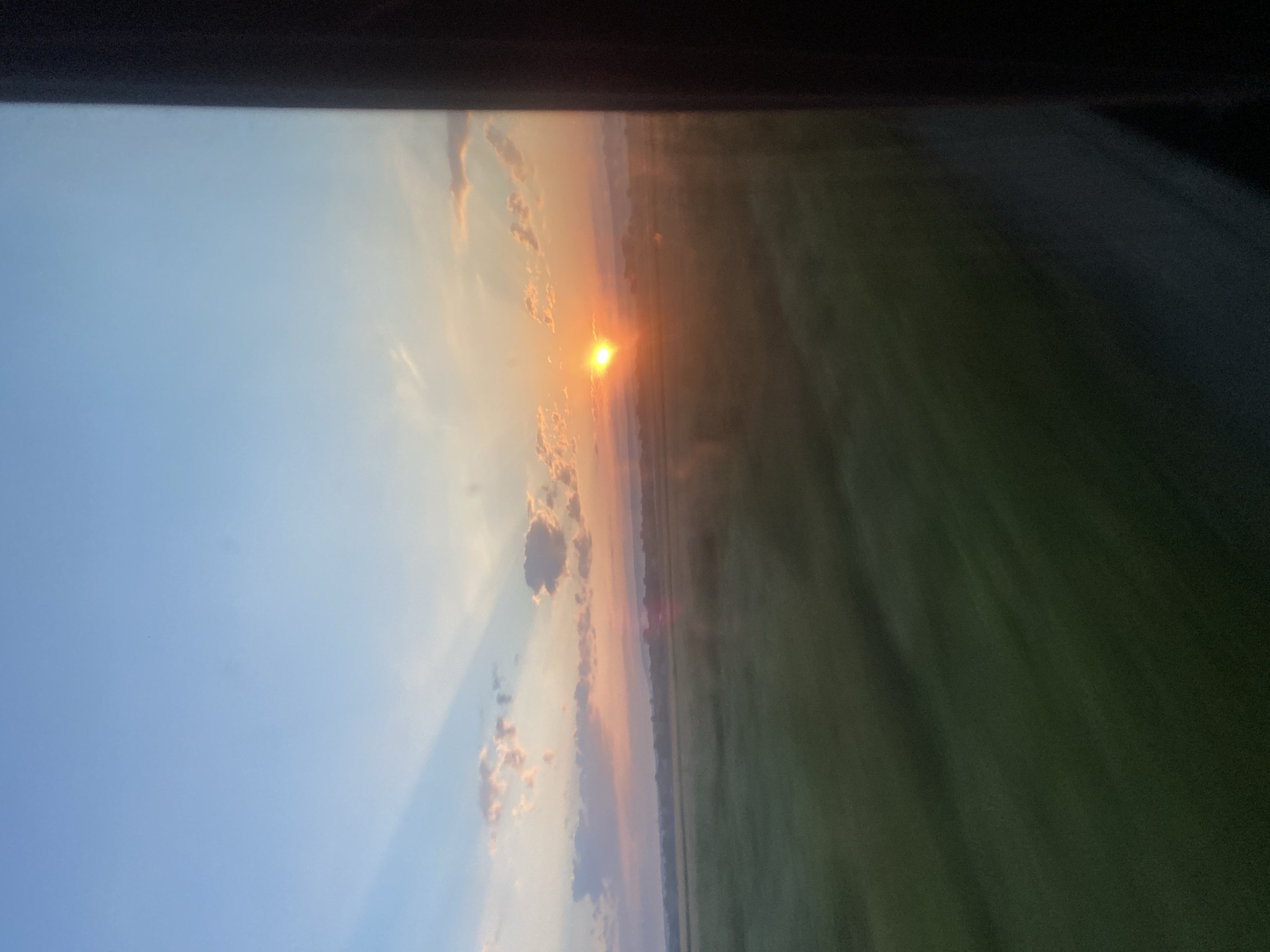 A photograph captures a sunset over a vast, green field in Novosiolovca, Moldova. The sun, a bright orange orb, is partially obscured by clouds and casts a warm glow across the sky, which transitions from a pale blue at the top to shades of pink and orange near the horizon. Wisps of clouds are scattered throughout the sky, some tinged with the colors of the sunset. The field below is a blur of green, suggesting movement, possibly from a vehicle or wind. The foreground is dominated by a dark, vertical structure on the right side of the image, which appears to be a window frame or a similar obstruction, partially obscuring the view of the landscape. The overall scene evokes a sense of natural beauty and tranquility as day transitions into night.