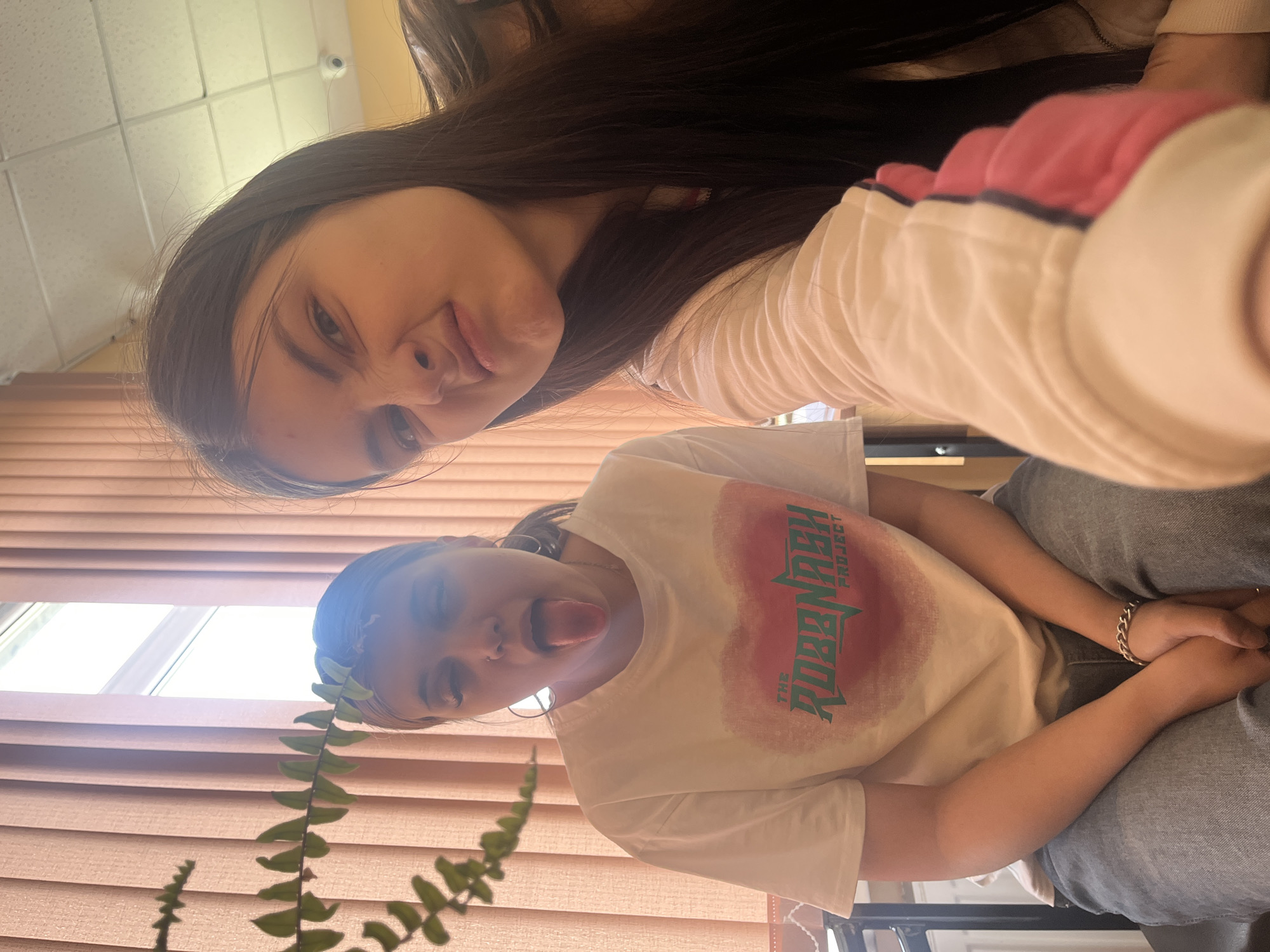 Two young women pose for a selfie in an indoor setting. The woman closer to the camera sticks her tongue out. A potted plant is visible in the foreground. The room appears to have window blinds. The location is not discernible as Saint Petersburg, Russia based on the image.