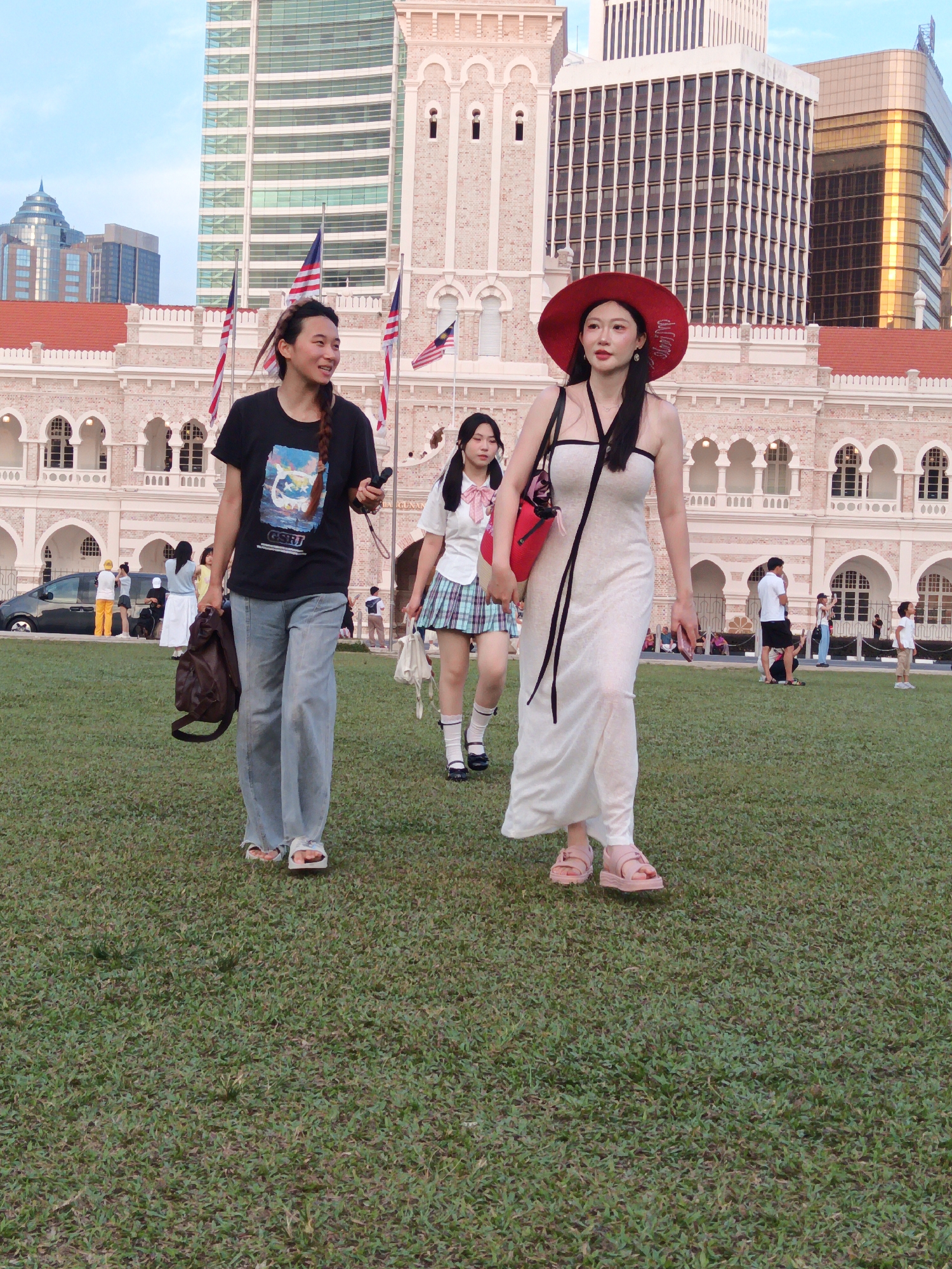 The image depicts the Dataran Merdeka in Kuala Lumpur, Malaysia, with the Sultan Abdul Samad Building as the primary backdrop. The building, characterized by its Mughal-inspired architecture and red-tiled roofs, stands prominently behind a lush green lawn. In the foreground, three young women are walking across the lawn.
The woman on the left, with her hair in a braid, wears a black t-shirt with a graphic print and light blue wide-leg jeans. She is carrying a brown bag in her left hand and holding a mobile phone in her right. To her right, a woman in a white collared shirt with a plaid skirt walks, holding a white bag. The woman in the center foreground is wearing a long, white, sleeveless dress and a wide-brimmed red hat. She is holding a red handbag over her shoulder and a mobile phone in her right hand.
In the mid-ground, other people are visible, including a group near a black van on the left and individuals sitting on the lawn further back. American flags are displayed on poles in front of the building. The sky is a clear blue, suggesting a pleasant day. The overall scene captures a casual, outdoor gathering in a historically significant urban space.