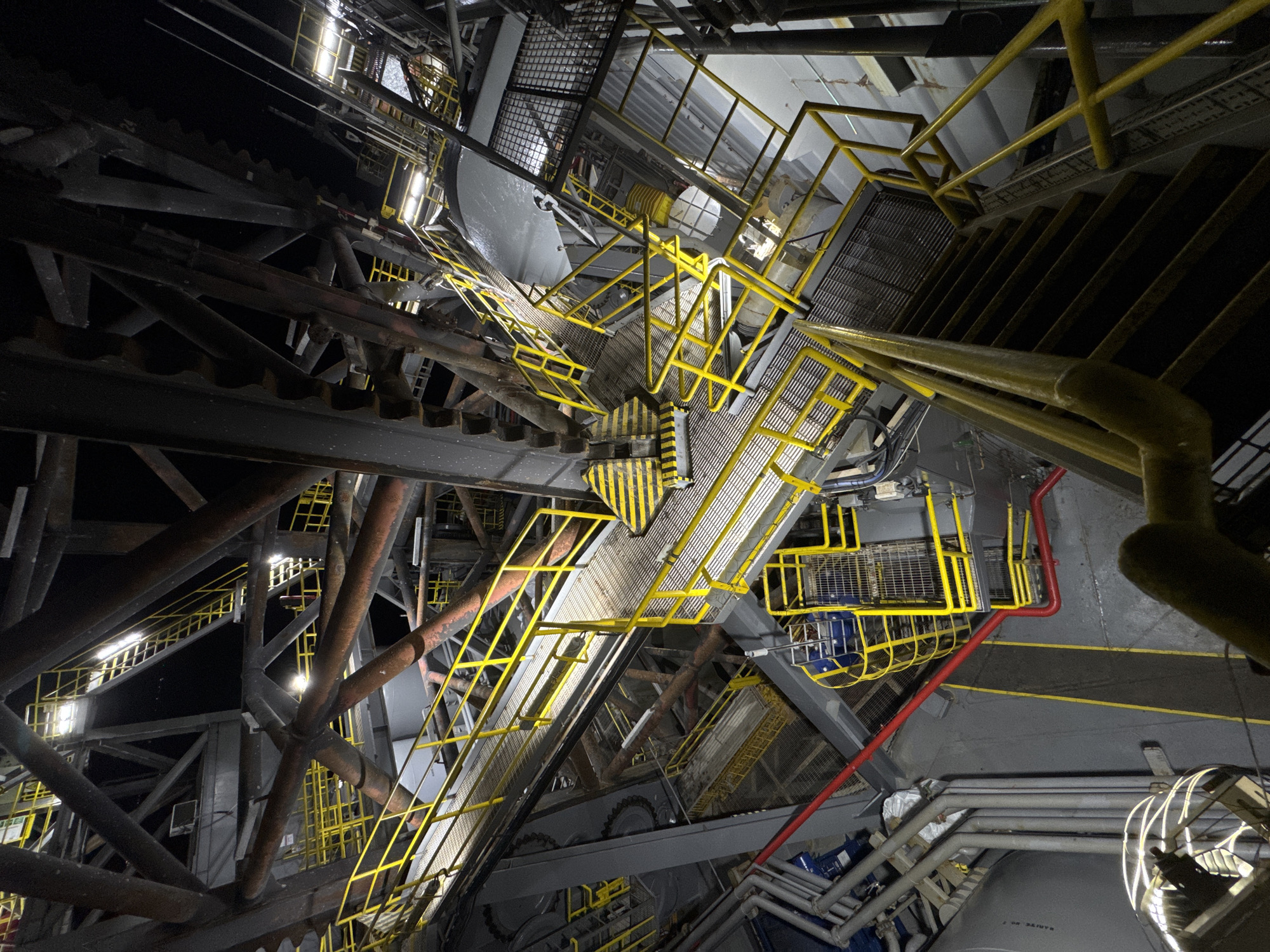 The image shows a complex industrial structure, possibly a drilling rig or a large piece of heavy machinery, viewed from a high vantage point looking downwards. The scene is dominated by an intricate network of metal beams, platforms, and staircases, many of which are painted bright yellow for safety. The lighting suggests it is nighttime, with artificial lights illuminating various sections of the structure. There are no people visible in the image. The environment appears to be an outdoor industrial setting, given the dark background and the vast scale of the structure. There is no identifiable context for a specific city or country.