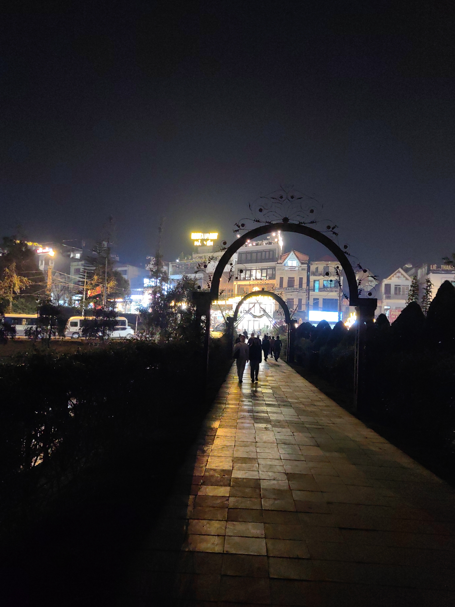 The image displays a nighttime scene in Sa Pa, Vietnam, featuring a stone-paved pedestrian walkway. The walkway, illuminated by artificial light sources, shows a warm, golden hue on its rectangular pavers. Approximately ten individuals, some silhouetted, are visible walking along this path, largely moving through two decorative metal archways ahead. The prominent archway, closer to the viewer, is adorned with intricate scrollwork at its apex. To the left of the walkway, a parallel street is visible, occupied by at least three vehicles, including a white passenger van, a white sedan, and a larger dark vehicle, partially obscured by dense dark foliage and coniferous trees. Beyond the archways and vegetation, a row of multi-story buildings line the street, illuminated by numerous windows and commercial signs in Vietnamese, such as "RESTAURANT ĐẶC SẢN HẢI YẾN" and "LIÊ THANH." The sky is dark, indicating a nocturnal setting, with ambient light emanating from the urban infrastructure.