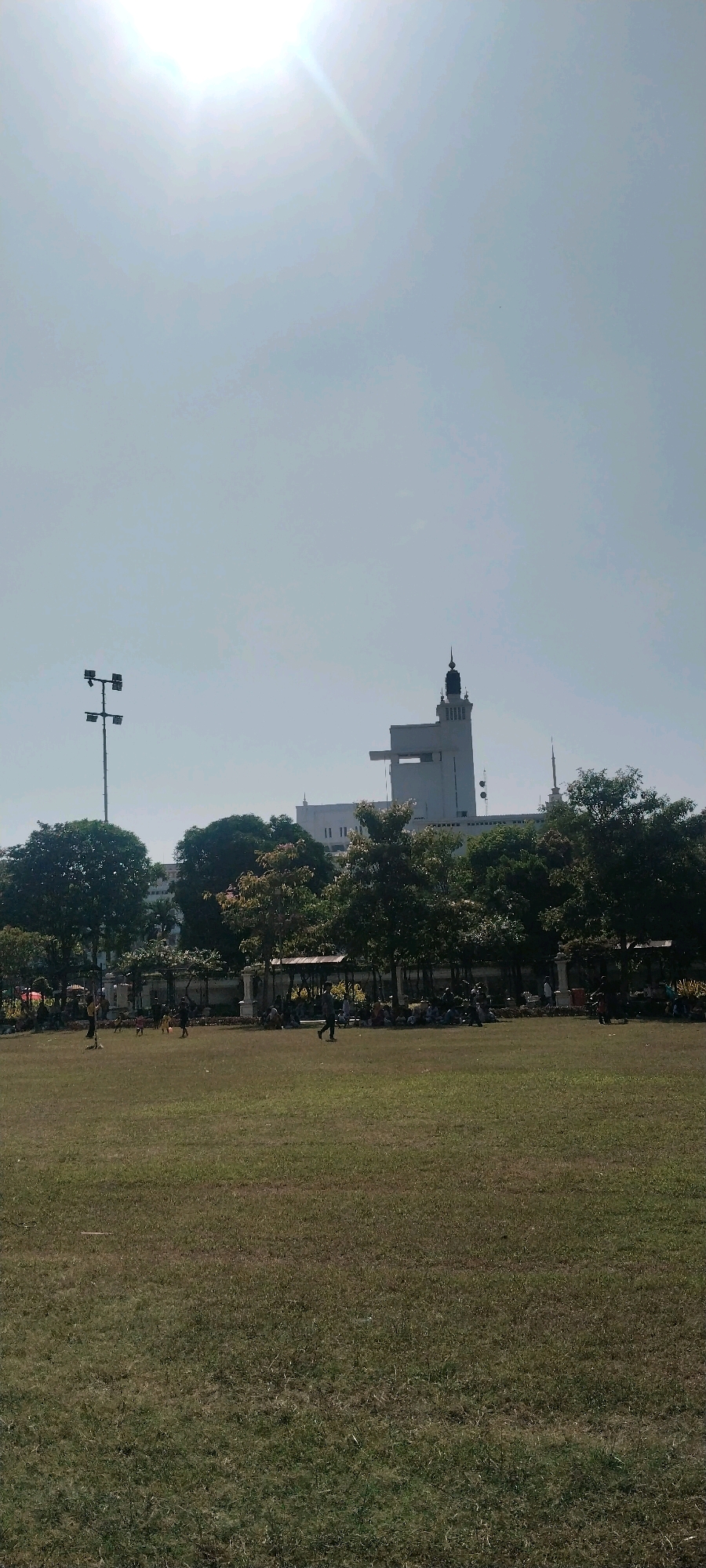A wide shot of a grassy field with a bright sky above shows a building in the distance. The building is white with a tall tower on its right side and a shorter rectangular structure to its left. The tower is topped with a dark dome. Trees with dark green leaves are in front of the building. In the foreground, people are scattered across the grassy field. Some are sitting, while others are standing and interacting. A light pole with four lights is visible on the left side of the image. The scene appears to be a public park in Surabaya, Indonesia.