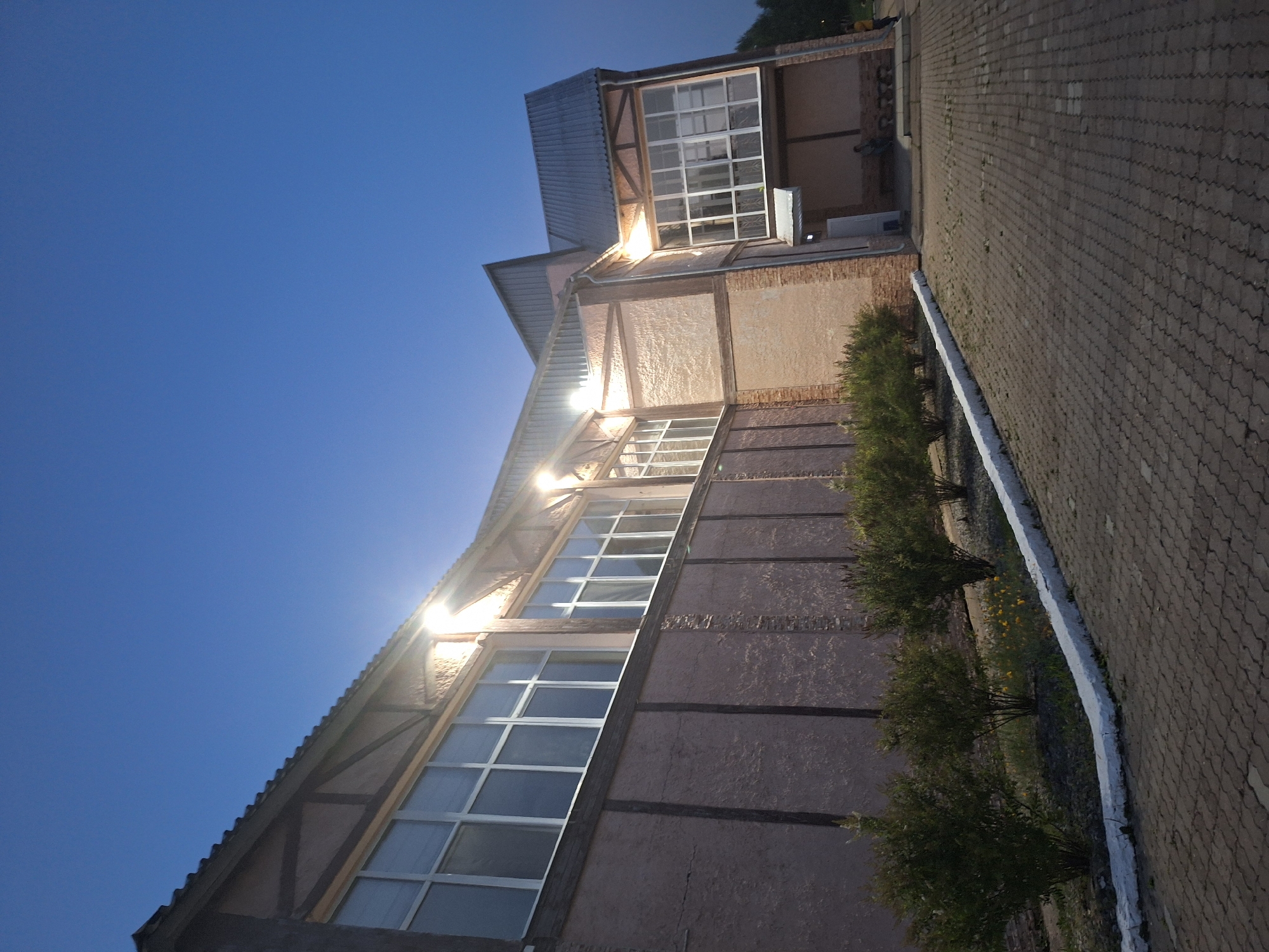 The image captures a building with a rustic architectural style, characterized by its textured, light-colored walls and dark wooden beams. The building features large, multi-paned windows that reflect the twilight sky. Outdoor lights illuminate the facade, casting a warm glow. In the foreground, a paved pathway made of interlocking bricks leads towards the building. A white-painted curb separates the pathway from a row of neatly trimmed bushes. Beyond the bushes, the ground is covered with grass and small yellow flowers. The sky above is a clear, deep blue, indicative of early evening. In the distance, at the entrance of the building, two individuals can be seen, one standing and another seated, suggesting the presence of activity or a gathering. The overall scene depicts an exterior view of a building in what appears to be a park or public area in Атамановка, Russia, during the twilight hours.
