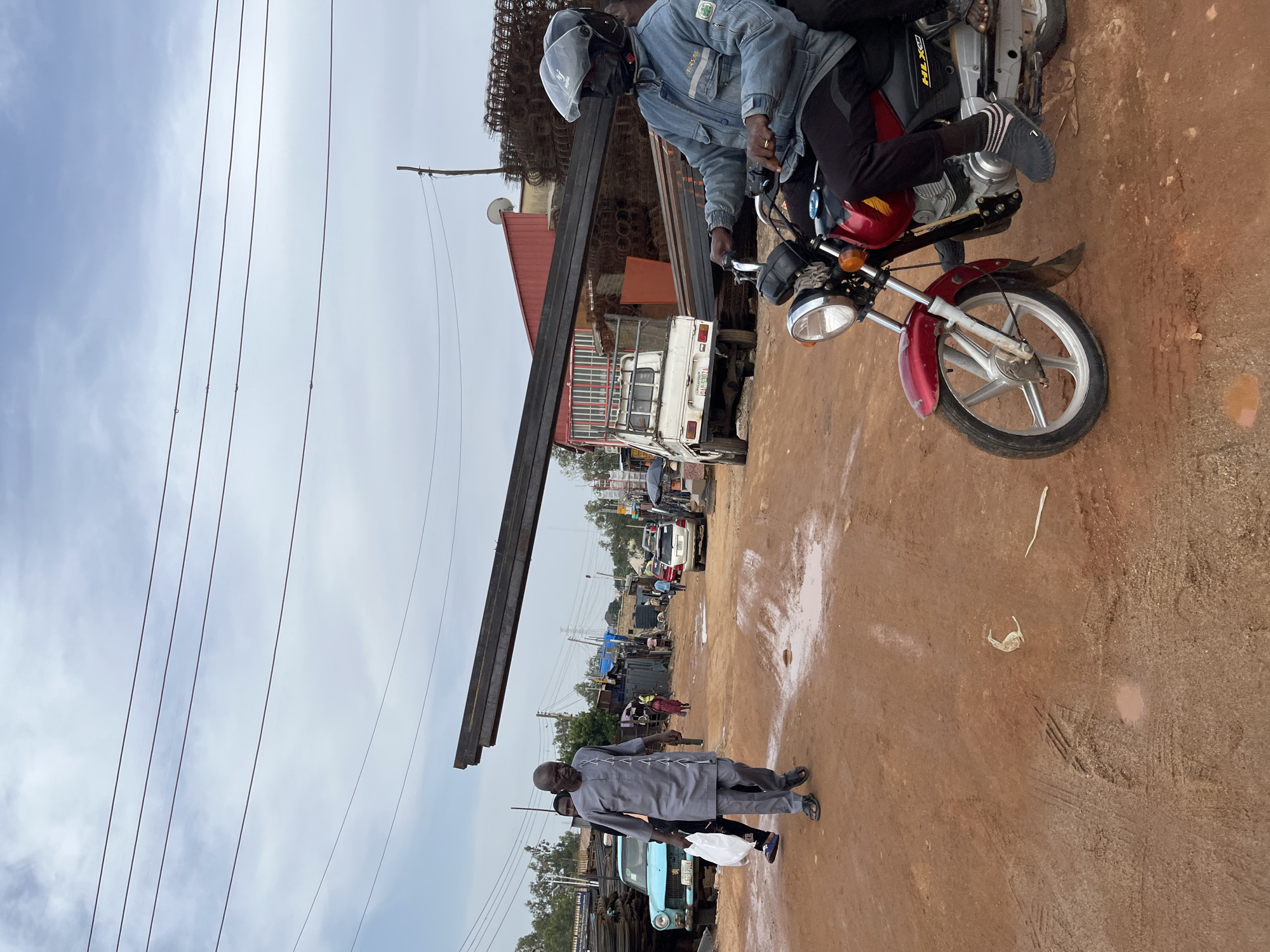 The image depicts a bustling street scene in Gura Top, Nigeria, likely during the daytime under an overcast sky. The focus is on a red motorcycle carrying two individuals. The rider, wearing a blue denim jacket and a helmet, is at the front, while a passenger in dark clothing sits behind. The motorcycle is positioned on a dirt road, which appears wet in places, suggesting recent rain.
In the background, a variety of objects and elements contribute to the sense of a commercial or industrial area. A tall, dark metal beam is prominently displayed, leaning diagonally across the frame. Behind it, a white truck with a license plate reading "LBS 3377" is visible, along with other vehicles and structures that suggest a marketplace or construction site. Several people are scattered throughout the scene, some walking and others gathered near vehicles.
Further in the distance, more buildings, trees, and utility poles can be seen, hinting at a somewhat developed but possibly informal urban environment. The presence of power lines strung across the sky further reinforces the notion of an inhabited area. A man in a grey traditional outfit walks in the foreground, holding a white plastic bag, adding another human element to the composition. The overall impression is one of everyday activity in a Nigerian town.
