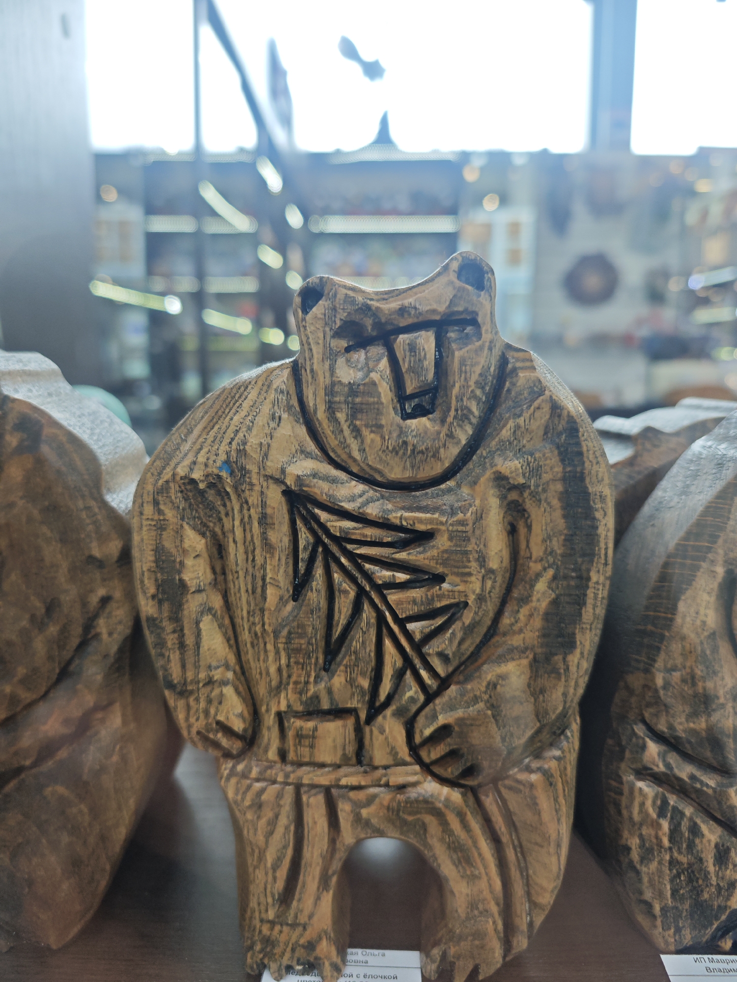 A close-up captures a carved wooden bear figurine, standing among others on a shelf within a shop. The bear features a stylized face and etched details, including a branch design on its chest. The setting is possibly a retail space in Bolshoye Savino, Russia, based on potential merchandise origins and signage at the bottom of the image, although text is illegible. The background shows blurry store displays, suggesting a general retail environment.