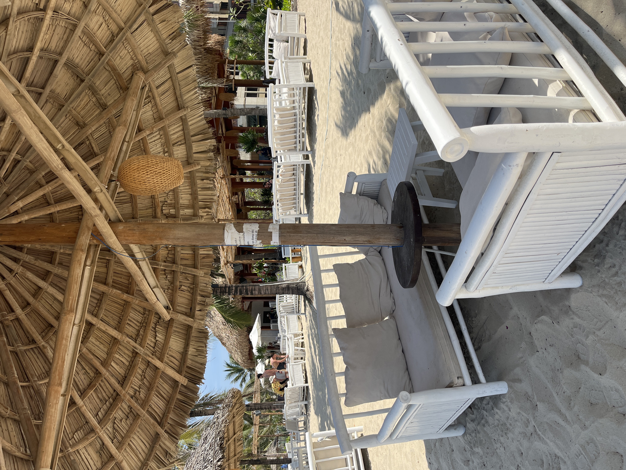 This image depicts a serene beach club or resort setting in Đà Nẵng, Vietnam, characterized by natural materials and a bright, sunny atmosphere.
The dominant feature in the foreground is a large, meticulously constructed thatched roof, serving as an umbrella or shelter. It's crafted from substantial bamboo poles forming the structural framework, overlaid with dried palm fronds or similar natural fibers. A distinctive woven spherical light fixture or decoration hangs from the bamboo framework.
Beneath this natural canopy, several white lounge chairs or daybeds with plush, light-colored cushions are arranged on the soft, sandy ground. These chairs also appear to be made from bamboo, painted white, contributing to a clean and inviting aesthetic. Small, dark wooden side tables are placed alongside some of the seating.
The sandy area extends into the background, revealing more white tables and chairs under additional smaller thatched structures and white umbrellas, suggesting dining or further relaxation zones. A few people are visible in the mid-ground, casually seated or walking, enjoying the beachside ambiance. Palm trees are scattered throughout the scene, enhancing the tropical feel, and modern buildings can be seen in the distant background.
The strong, clear shadows cast on the sand indicate bright daylight, likely late morning to early afternoon, under a clear sky. A partially visible sign or paper is affixed to one of the wooden support posts, with some text discernible, including the word "under" and possibly "please". The overall scene conveys a strong sense of leisure, comfort, and vacation.