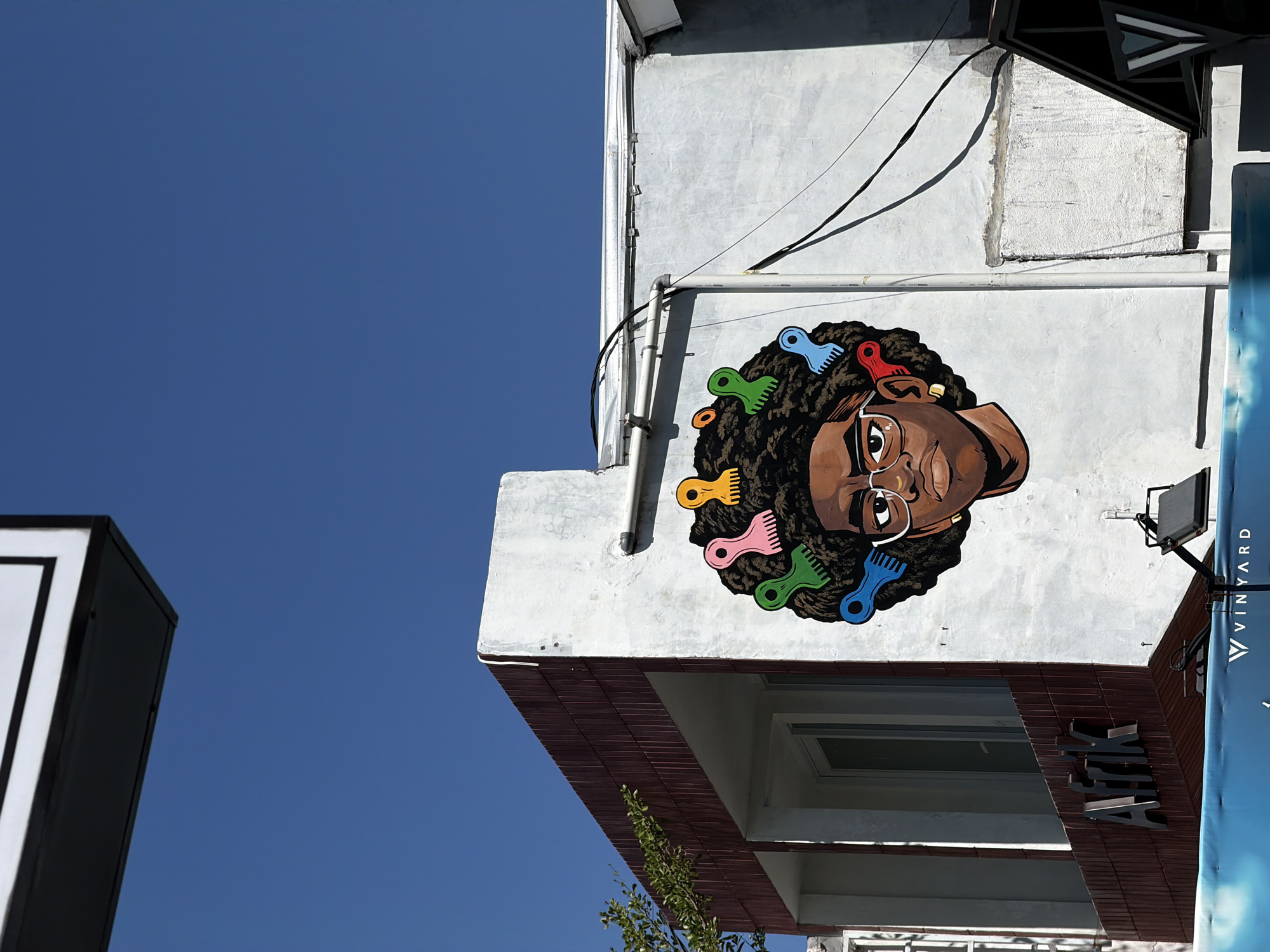 The photograph depicts a mural on the side of a building in North Kuta, Indonesia. The mural features a stylized image of a woman's head with an afro hairstyle adorned with colorful hair combs. The woman is depicted with glasses and an earring. The mural is painted on a white building with a reddish-brown tiled overhang. A window is visible beneath the mural. A sign with the name "VINYARD" is partially visible on the right side of the image. The sky is a clear blue. The photograph is taken from a low angle, looking up at the building. Another building is slightly visible at the left.