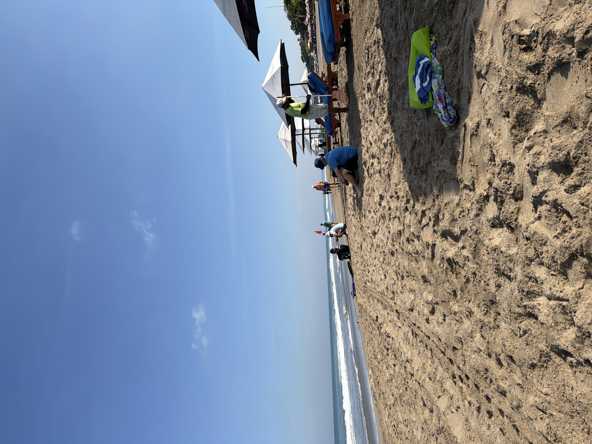 A sunny daytime scene unfolds on a sandy beach in Kuta, Indonesia. The image is rotated 90 degrees clockwise, presenting a vast expanse of bright blue sky with a few wispy clouds dominating the upper left, while the light brown sand covers the lower right. The horizon line is formed by the ocean, with gentle white waves lapping at the shore.
On the sandy beach, various activities are taking place. In the foreground, a neon green beach bag or towel is laid out next to a colorful sarong and blue flip-flops. Further along the beach, several people are visible. One individual, wearing a blue shirt and a dark hat, is kneeling or crouching on the sand, possibly engaging in sand play or digging. Another person, dressed in a light green shirt and a light hat, stands near a white beach umbrella, seemingly setting it up or adjusting it. Other beachgoers are scattered further down the shore, near more umbrellas and blue lounge chairs, suggesting relaxation or leisure. A small red flag is visible near the water's edge.
The beach is lined with multiple umbrellas, some white and some black, providing shade. Text is visible on some of the white umbrellas, including "BALI PARADISE", "SINGAPORE", and "TROPICAL". The bright sunlight casts distinct shadows on the textured sand, indicating a clear, pleasant day, likely in the late morning or early afternoon. The overall atmosphere conveys a typical tourist beach setting, with people enjoying the sun and sea.