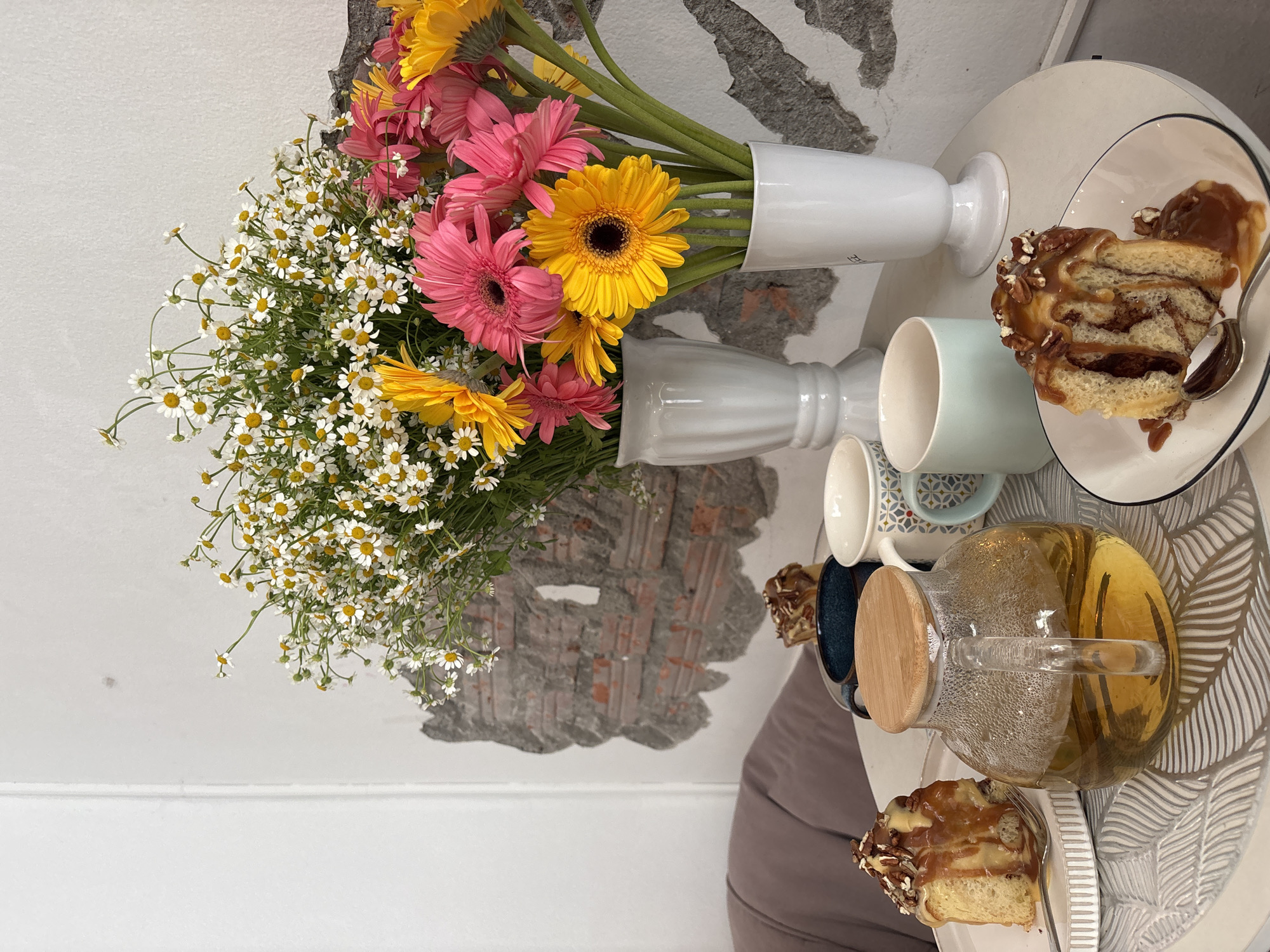 The image depicts a table setting, possibly in a café or home setting in Phuket City Municipality, Thailand.
Subjects/Objects: A large bouquet of mixed flowers, including chamomile and gerbera daisies, in two white vases. A white table with a modern leaf pattern, holding a clear glass teapot filled with a yellow liquid (likely tea). Two cups are present, one light blue and one patterned. Two plates hold slices of a caramel and pecan-topped pastry. A spoon rests on one plate.
Environment: The table is set against a white wall, with a patch of exposed brickwork.
Action: The scene suggests a tea or coffee break, with the food and beverages laid out.
Interactions: There is no observable interaction, as the scene is static. The arrangement implies an intention to share the food and beverages.
Details: The pastries are dripping with caramel sauce, suggesting a sweet and indulgent treat. The presence of fresh flowers adds a decorative and pleasant touch to the scene. The exposed brickwork gives a rustic or modern-industrial feel to the interior. The overall composition suggests a relaxing and aesthetically pleasing moment.