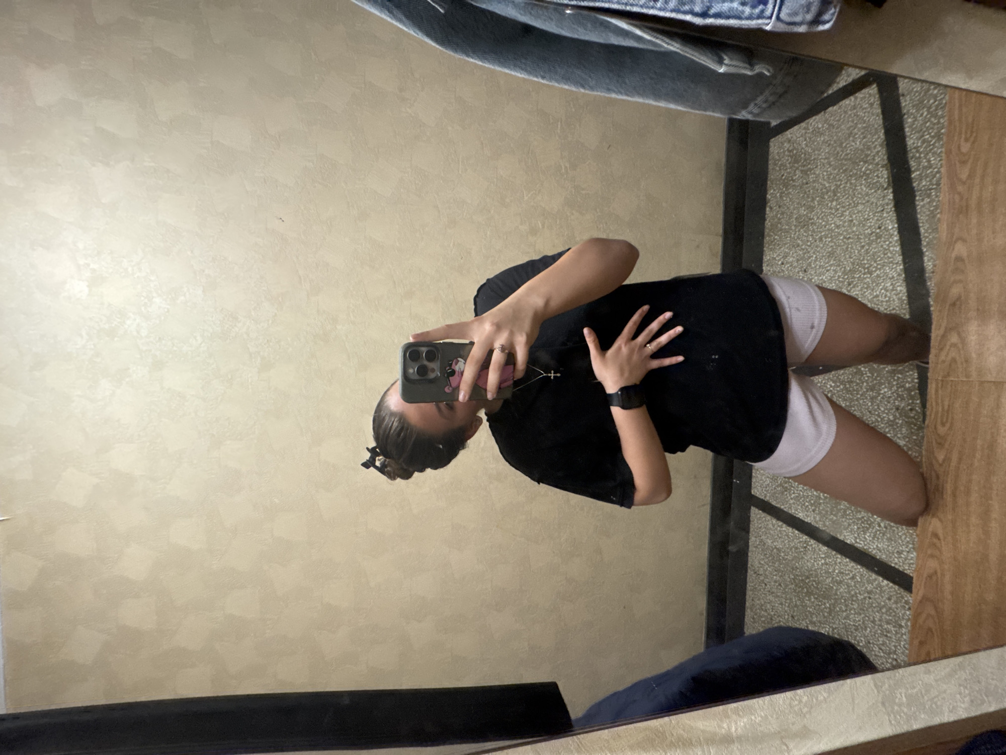 A person is captured in a mirror selfie, standing in an indoor setting. The image is rotated 90 degrees clockwise. The individual, appearing to be female, is wearing a black t-shirt, white shorts, and has their dark hair tied back with a clip. They are holding a smartphone (an iPhone with a visible camera module) with their right hand to take the photo, while their left hand rests on their torso. Notable accessories include rings on fingers, a black smartwatch on the left wrist, and a silver cross necklace.
The mirror reflects a light beige wall with a subtle textured pattern behind the person. To the right of the person's reflection (viewer's left), a dark denim fabric, likely clothing, is visible. Below the person's reflection, a gray, speckled floor is seen, along with a dark structural element (possibly a frame or beam). On the far right of the reflection, a wooden surface suggests part of the room's furnishing. The overall impression is an ordinary room, possibly in Sofia, Bulgaria, given the provided location context. There is no clearly legible text visible in the image.