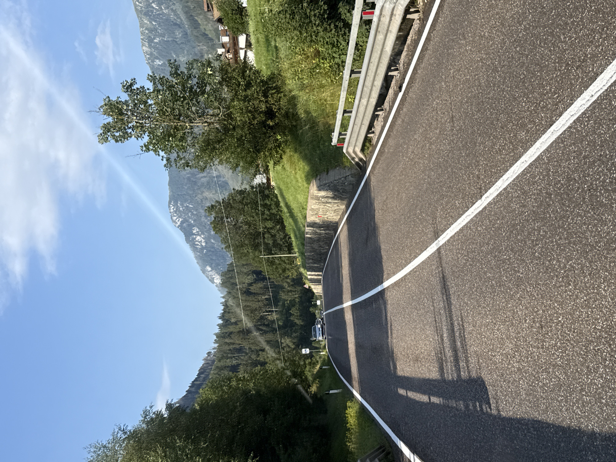 The image depicts a scenic asphalt road curving through a mountainous landscape, likely in or near Moena, Italy, given the context. The road is marked with white lines, and shadows of trees are cast upon its surface. A silver car is visible in the distance, traveling on the road. On the right side of the road, a metal guardrail lines the edge, and a stone retaining wall is present on the left. Lush green trees and grass cover the slopes leading up to the mountains in the background, which are partially snow-capped. The sky is a clear blue with a few wispy clouds, and a faint rainbow-like arc is visible in the upper portion of the frame. In the upper right corner, buildings with tiled roofs can be seen nestled on a hillside. The overall scene suggests a pleasant, sunny day in a mountainous region.