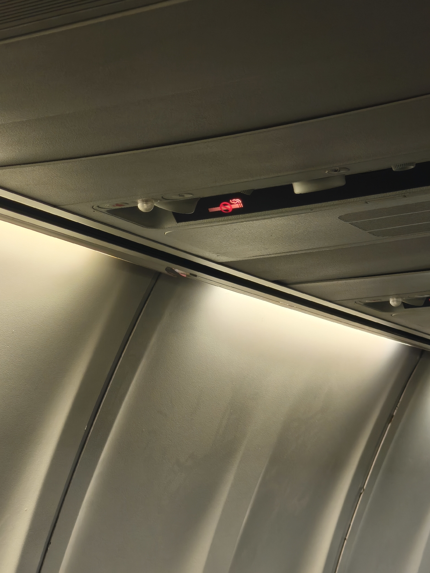 The image is a low-angle, interior shot of an airplane cabin. The ceiling panels are visible, with a prominent "no smoking" sign illuminated in red. The sign features a cigarette with a red circle and slash through it. Below the ceiling, the curved wall of the cabin is visible, with recessed lighting creating a soft glow. The overall impression is one of a typical airplane interior, likely in flight. The image does not provide any contextual information that would place it in Kuta, Indonesia, or any specific city. There are no people or any discernible actions taking place. The scene is static and focused on the cabin's infrastructure.