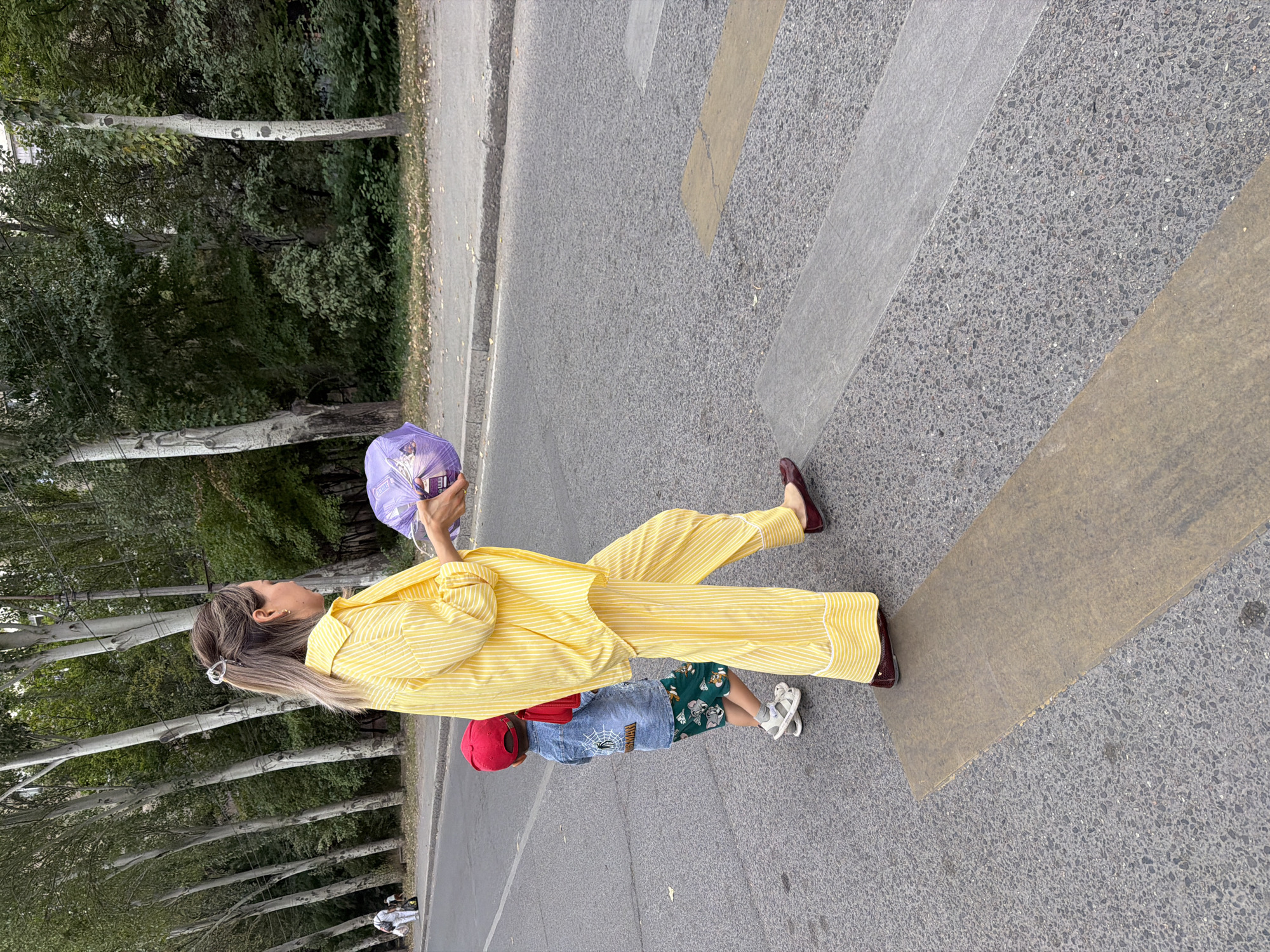 A woman and a young boy are crossing a street at a pedestrian crossing. The woman, wearing a yellow striped pajama-like outfit and dark red shoes, is holding a purple balloon. The boy, in a denim jacket, green shorts, and a red hat, walks beside her. They are on a paved road with trees lining the background. The overall scene suggests a casual outdoor setting in a city.