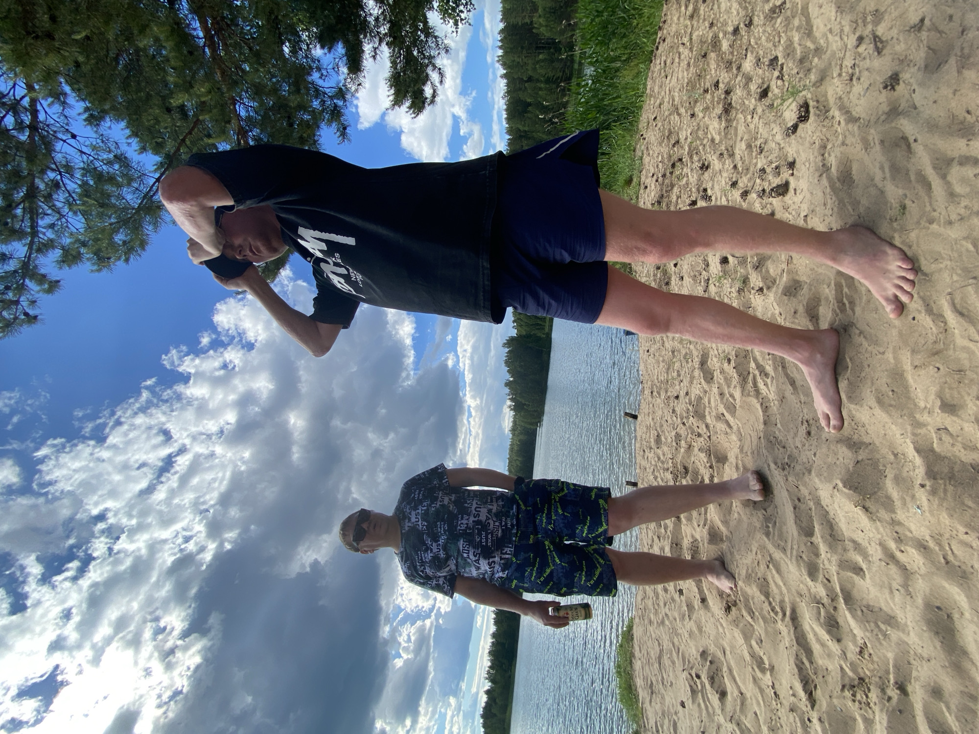 Two men stand on a sandy shore near a body of water, possibly a lake, in Novyi, Russia. The man on the left wears a patterned t-shirt and patterned shorts, holding a can in his right hand. He is also wearing sunglasses. The man on the right is wearing a black t-shirt and dark blue shorts and has his hands near his head, adjusting a dark cap. The sky is partly cloudy with blue patches and the surrounding area is covered with green trees. The ground is sandy with pine cones scattered on it.