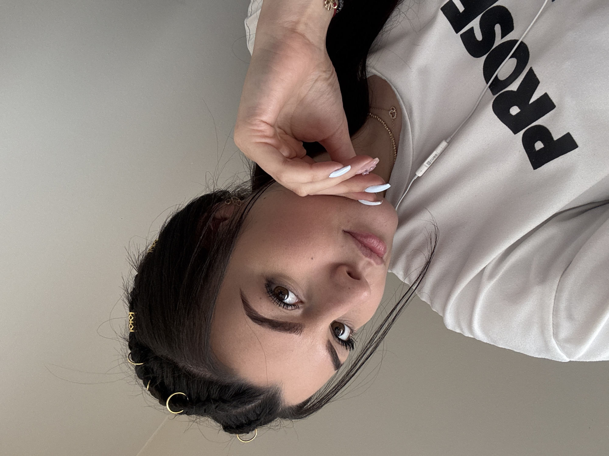 A close-up, eye-level indoor portrait captures a young female with dark hair and brown eyes, facing slightly to her left (viewer's right) with her head tilted. Her dark hair features two braids on the left side, adorned with small gold rings and one gold geometric charm. Loose strands of hair frame her face. She has visible eyelashes, defined eyebrows, and a small mole on her upper right cheek. Her fingernails are long and almond-shaped, painted pale blue on most fingers, with the ring finger of her right hand (viewer's left) showing a translucent pink or clear polish with a subtle design.
She is wearing a white long-sleeved shirt with the partially visible black block letters "PROSE" across the chest. A delicate gold necklace with two interlocking heart pendants is around her neck, and small gold earrings are visible. A red and black bracelet with a gold clover-like charm is on her left wrist. Her right hand is positioned with fingers gently touching her chin and cheek. White wired earphones, with a control module labeled "cube," hang down the front of her shirt. The background is a plain, light gray wall, indicating an indoor setting. There are no visual cues within the image to specify a location in Didim, Turkey.