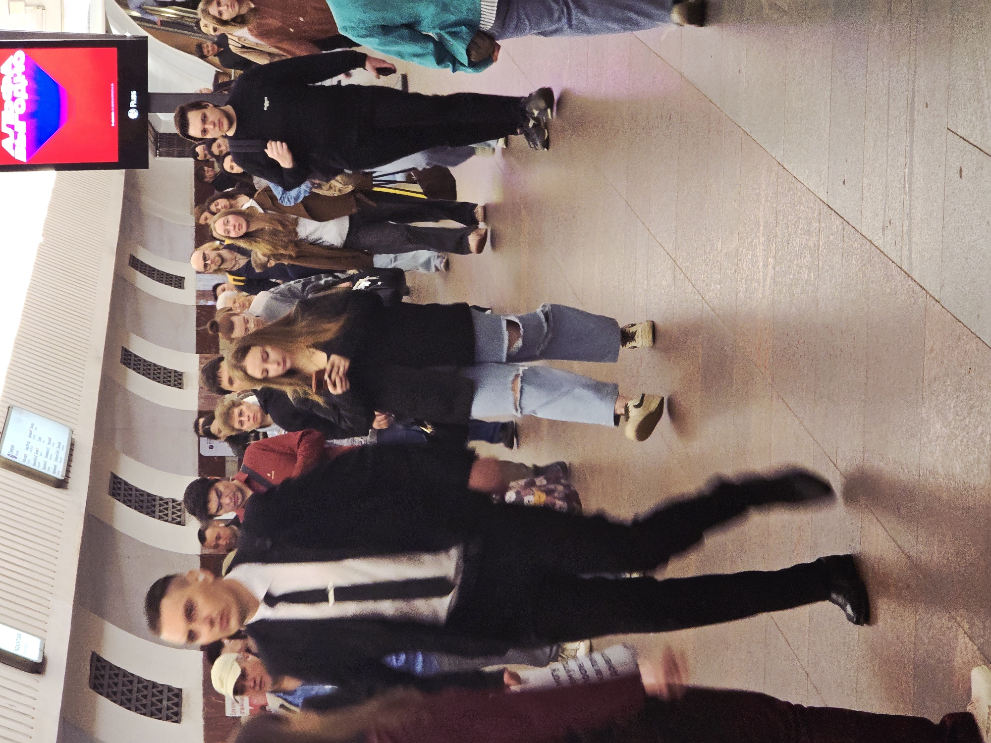 The image shows a group of people, mostly young adults, standing or walking in what appears to be an underground transportation hub, possibly a subway or train station in Unknown, Unknown. The setting features a patterned wall with air vents, and digital display boards, one of which displays an advertisement with red and blue graphics. The people are dressed in casual clothing, with some individuals in jackets or suits. A person wearing a black suit with a white stripe seems to be in motion. The scene suggests a busy public transportation area with people waiting or transiting through the location.