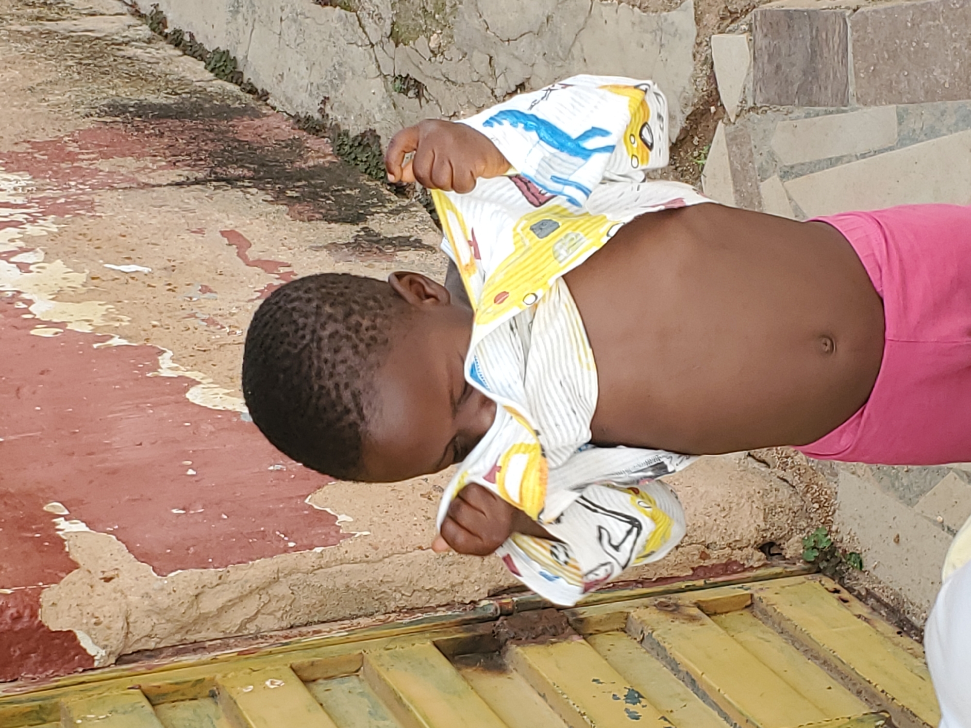 A child is shown from the waist up, lying on their side. They are wearing a white shirt with colorful patterns of yellow and blue drawings, possibly depicting cars or other objects. The child's midriff is exposed, and they are wearing pink shorts. Their head is turned downwards, and their face is not fully visible. The child's hands are near their chest, holding the shirt. The background consists of rough, textured ground and a wall made of various stones and concrete. Some green plants are growing in the cracks between the stones. The ground appears to be a mixture of red and beige tones, with patches of wear and tear. A yellow metal structure, possibly a grate or a door, is visible at the bottom of the image. The lighting suggests it is daytime, and the overall scene appears to be outdoors, possibly in an urban or residential area.