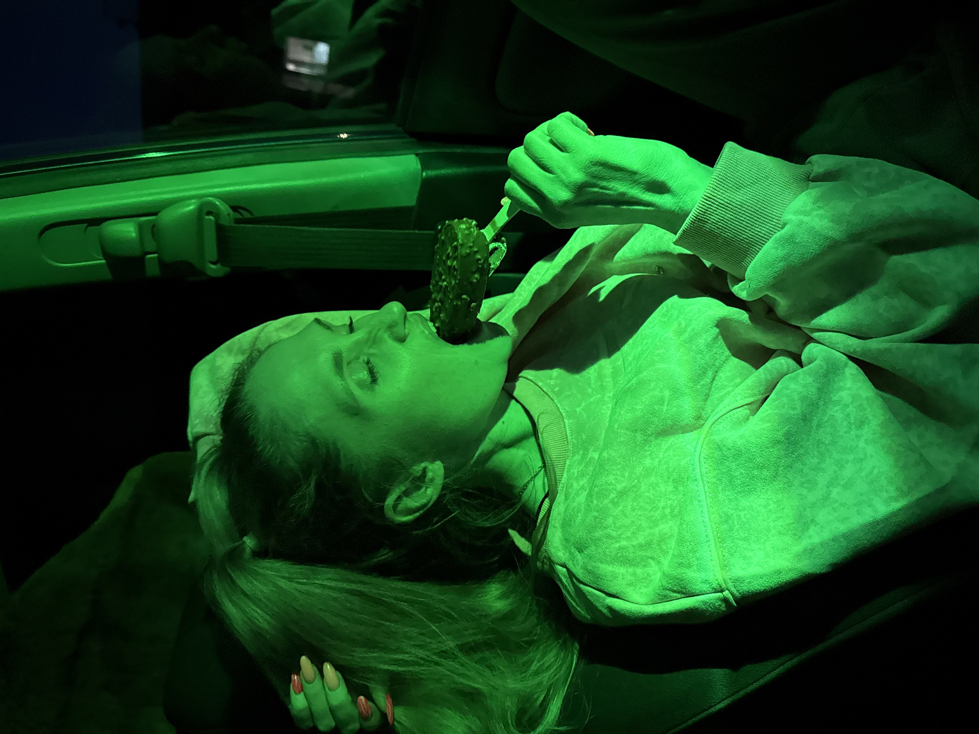 A woman with long blonde hair is lying down in what appears to be a vehicle, possibly a car, illuminated by a bright green light. She is wearing a sweatshirt. She is holding a green object on a stick up to her mouth. A seatbelt and the interior of the vehicle are visible. The image has a dark background. The location, Chaikovsky, Russia, is not verifiable within the confines of the given image.