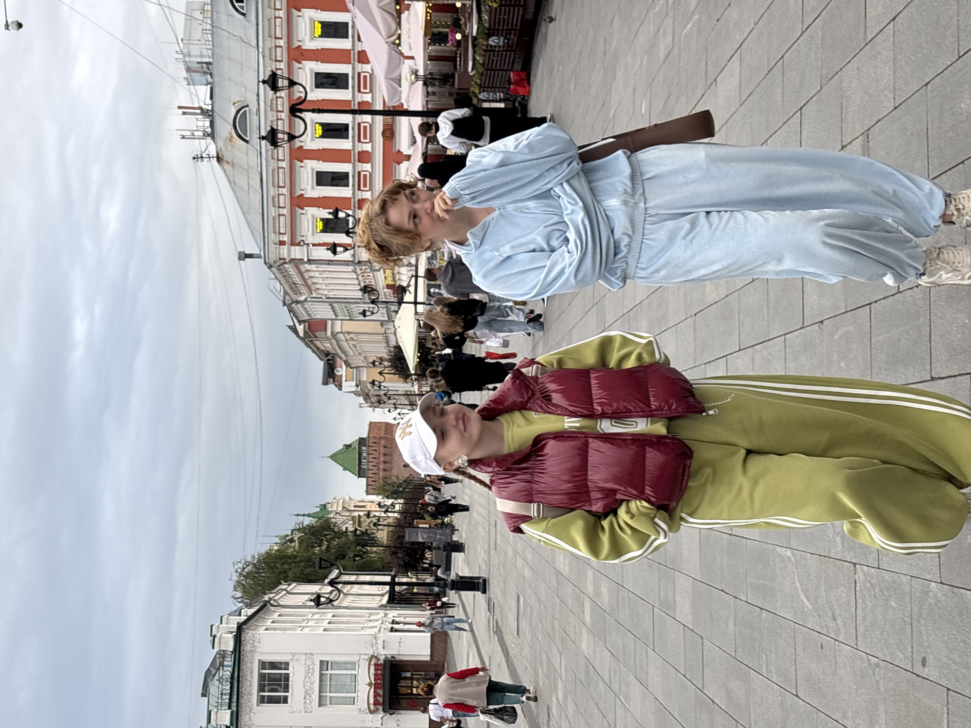 Two young women walk down a cobblestone street in Nizhny Novgorod, Russia. The woman on the left wears a lime green tracksuit with white stripes down the legs and a burgundy puffer vest. She has a white baseball cap on and a backpack slung over one shoulder. The woman on the right wears a light blue velour tracksuit and has her hand near her mouth, looking to the side. In the background, several people walk in both directions. The street is lined with buildings, some of which have red and white facades. In the distance, a green-roofed tower is visible. The sky is overcast.