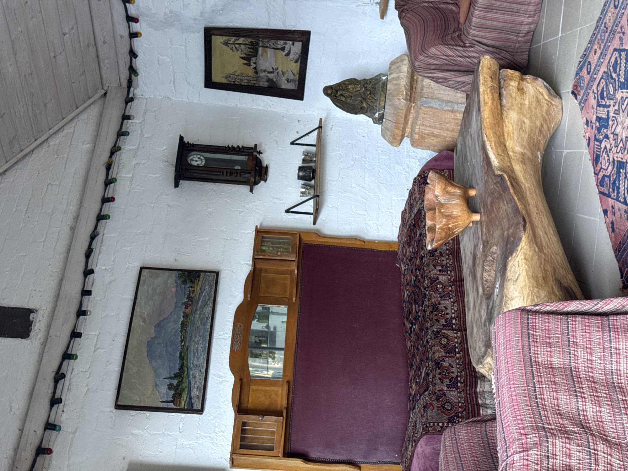 The image depicts an interior scene, likely a living room, filled with rustic and vintage decor. Dominating the foreground is a large, organic-shaped wooden coffee table with a rough-hewn texture. On this table sits a carved wooden bowl. To the left, partially visible, is an armchair draped with a pink and striped textile. Behind the armchair is a bed with a deep red headboard, covered by an intricately patterned rug.
The background reveals a white brick wall adorned with several decorative elements. A large, framed landscape painting hangs on the wall above the bed, depicting a mountain range, a river, and a village. To the right of this painting, a dark wooden grandfather clock with a visible clock face is mounted on the wall. Below the clock, a small wooden shelf holds several small metallic cups or votives. Further to the right, another framed painting, this one depicting a snowy landscape with trees, is displayed. Below this painting, a bronze sculpture of a hooded figure holding a child rests on a multi-tiered wooden pedestal.
Decorative string lights with colored bulbs are strung along the upper part of the white wall and across the angled ceiling, adding a festive or ambient touch. The flooring appears to be tiled. The overall impression is one of a cozy, eclectic space with a focus on natural materials and antique pieces. There is no discernible connection to Alagir, Russia, in the image itself; the description is purely based on the visual elements present.