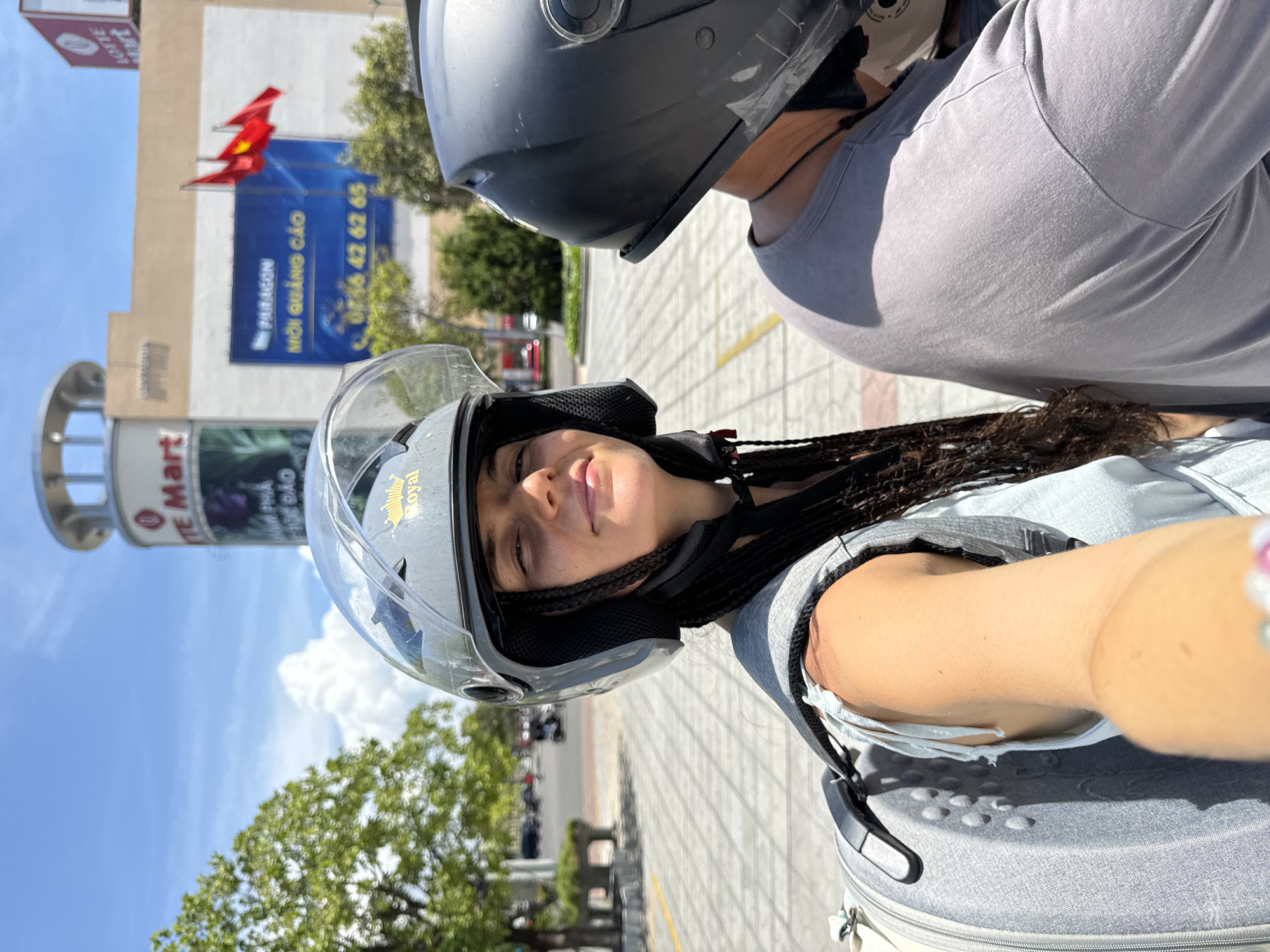 In Phu Thuy Ward, Vietnam, on a bright, sunny day with scattered clouds, a young woman with dark braided hair, wearing a light blue shirt and a grey helmet, is taking a selfie from the back of a motorbike. She is positioned in the foreground, her face partially visible as she looks towards the camera. A backpack is slung over her shoulders. To her right, another person wearing a grey t-shirt and a black helmet is also on the motorbike, facing forward. In the background, a large building with "Lotte Mart" and "Paragon" signage is visible. Several Vietnamese flags are displayed on poles in front of the building. The street in front of them is paved with interlocking bricks, and there are trees and other motorbikes in the distance.