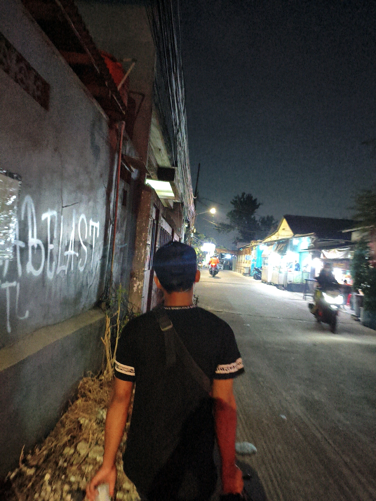 The image depicts a nighttime street scene in Bekasi, Indonesia, viewed from behind a person. The foreground shows the back of a person wearing a black t-shirt with white bands on the sleeves and a dark cap. They are holding a clear plastic bottle in their right hand. To the left, a rough concrete wall is covered in white graffiti. To the right, the street is paved and illuminated by various light sources from businesses lining the road. Several motorcycles are visible on the street; one rider is seen in the distance on the left, and another is in motion on the right. The buildings appear to be simple structures with open fronts, possibly food stalls or small shops. Overhead, a dense network of electrical wires is prominent against the dark sky. The overall impression is that of a busy, working-class urban environment at night.