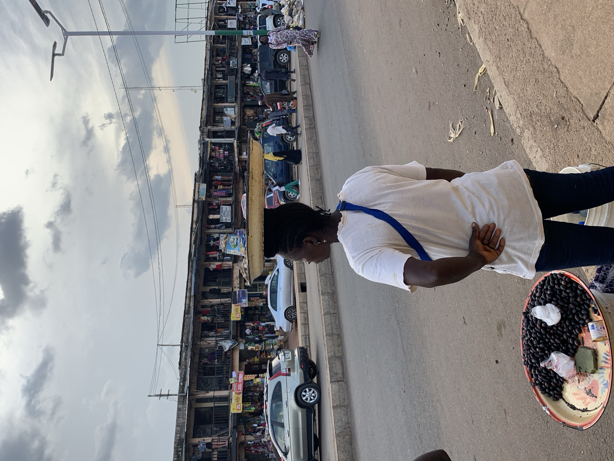 A woman, dressed in a white t-shirt and dark pants, stands with her back to the camera, facing a busy street scene. She is wearing a blue strap across her shoulder. In front of her, to the right, is a large, oval tray filled with dark, round fruits, possibly olives, along with a small tin and some wrapped items. The tray is placed on the asphalt ground.
The background reveals a vibrant street with multiple stories of shops and stalls lining the left side of the road. The shops are packed with various goods, and people are visible in and around them, suggesting a bustling market environment. Several cars are parked along the roadside or driving by, including a white sedan and a silver SUV. Power lines crisscross above the street, and a utility pole with lights is also visible.
The sky is overcast with dramatic clouds, indicating a possibility of rain or a generally cloudy day. The overall scene suggests a typical day in a busy urban market in Jos, Nigeria, characterized by commercial activity and street life. The woman appears to be a vendor, perhaps selling the fruits from her tray.