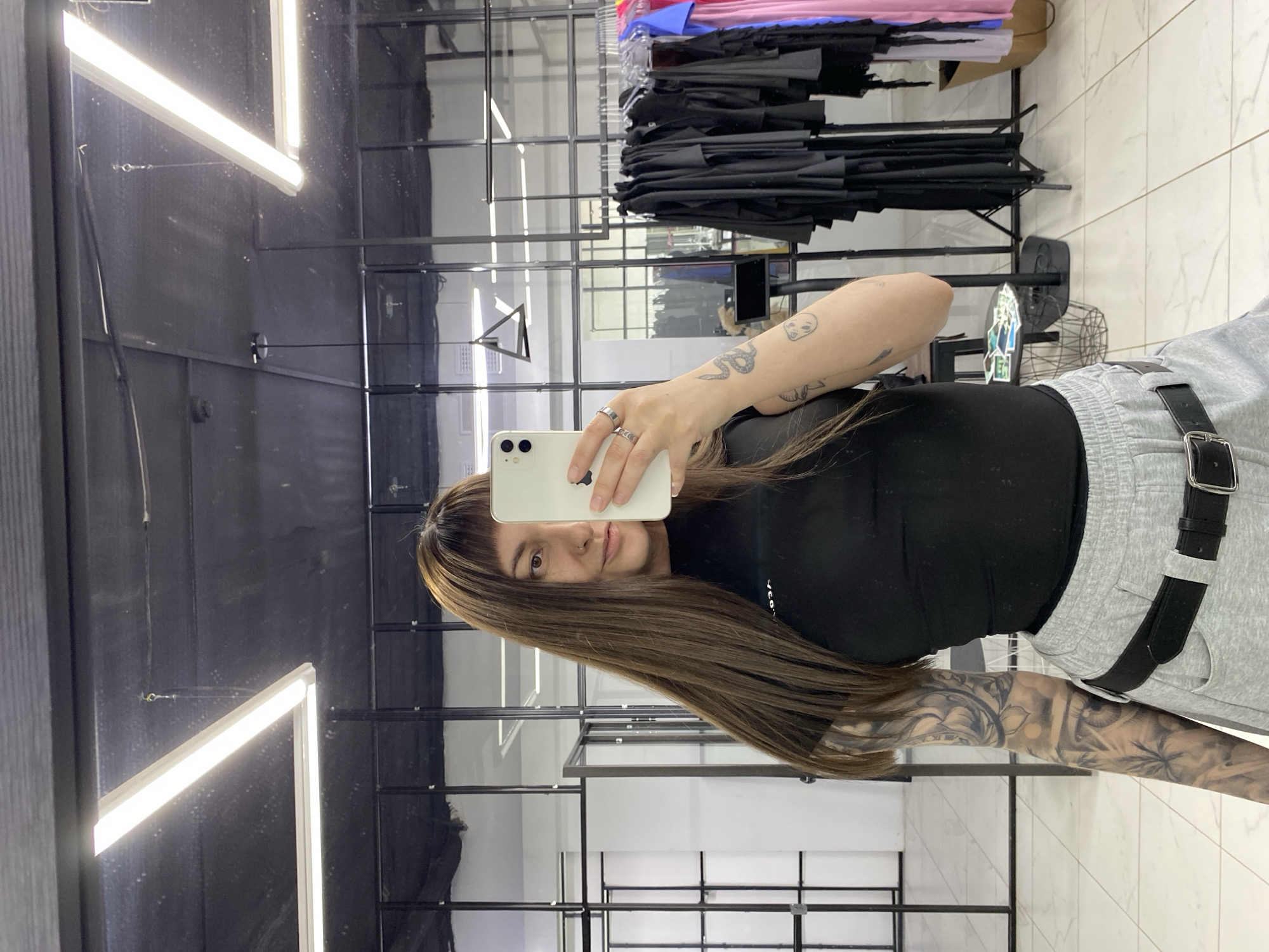 A woman with light brown hair and tattoos on her left arm is taking a selfie in a store. She is wearing a black top and gray sweatpants with a black belt. She is holding a white iPhone in her right hand. The store appears to be selling clothing, with racks of clothes visible in the background. There are also fluorescent lights on the ceiling. The location is likely a clothing store in Votkinsk, Russia.