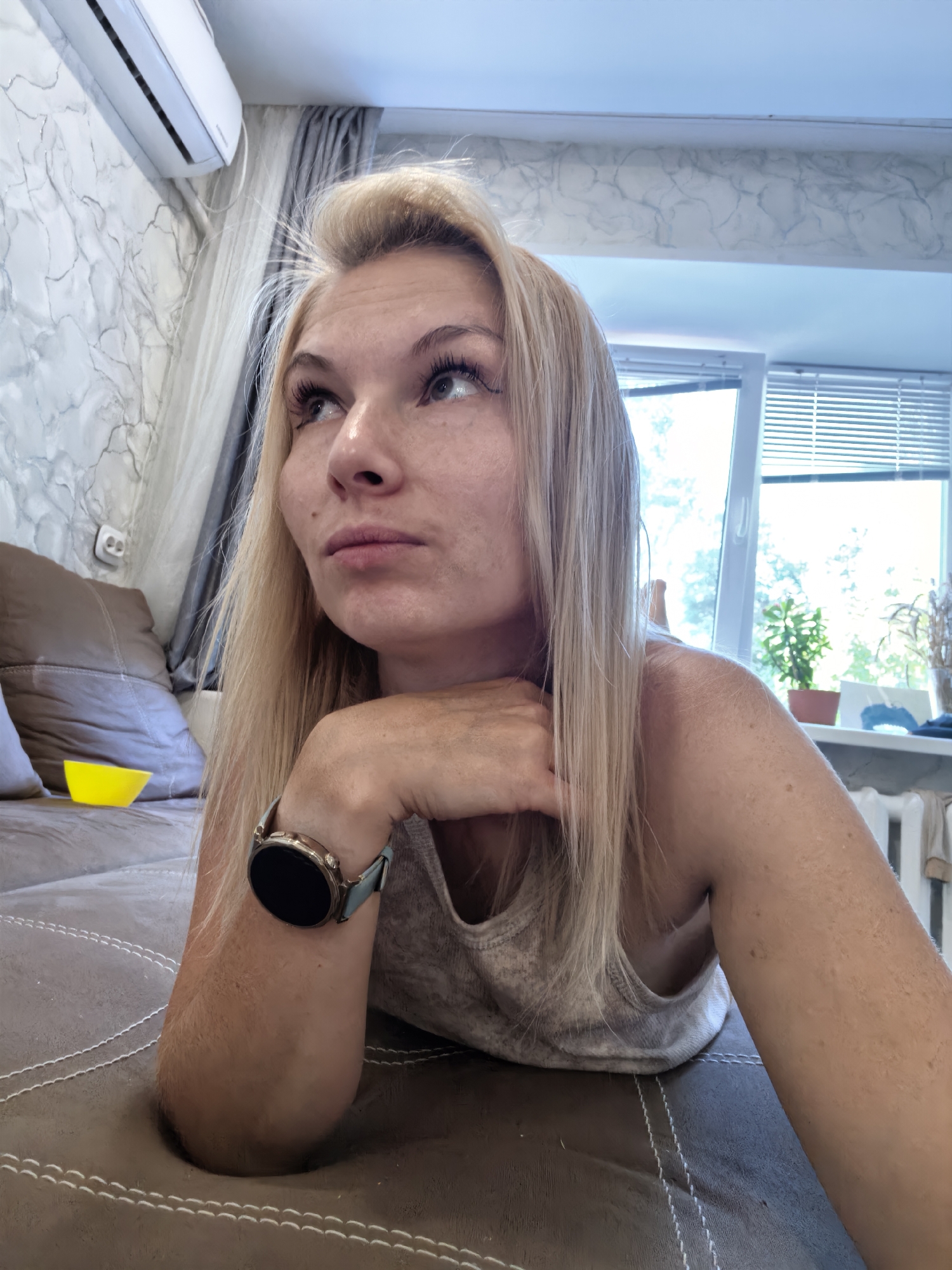 A blonde woman lies on a sofa with her head propped on her hand. She is looking to her left and up. She is wearing a light-colored tank top and a watch on her left wrist. A yellow bowl is visible on the sofa next to her. The room has an air conditioner mounted on the wall, a window with blinds, and some plants on the windowsill. The sofa appears to be made of brown leather. The walls are decorated with a textured wallpaper.